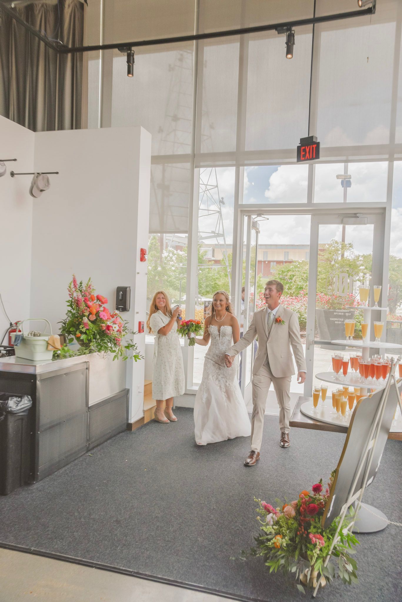 Newlyweds walk hand-in-hand through a doorway, followed by a bridesmaid applauding. Wedding reception with flowers and drinks.