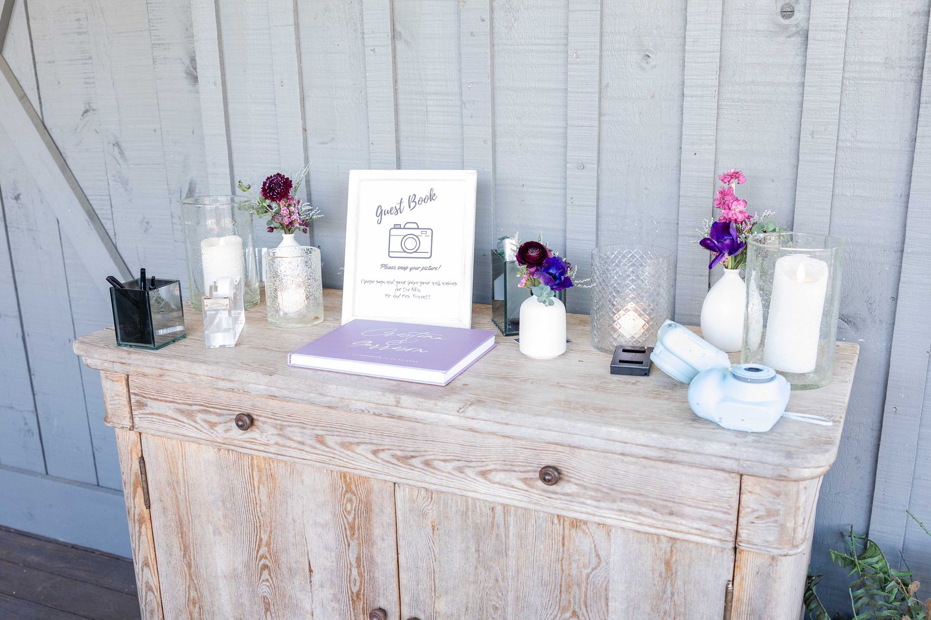 A rustic wooden cabinet displays a guest book, flowers, candles, and a sign with calligraphy.