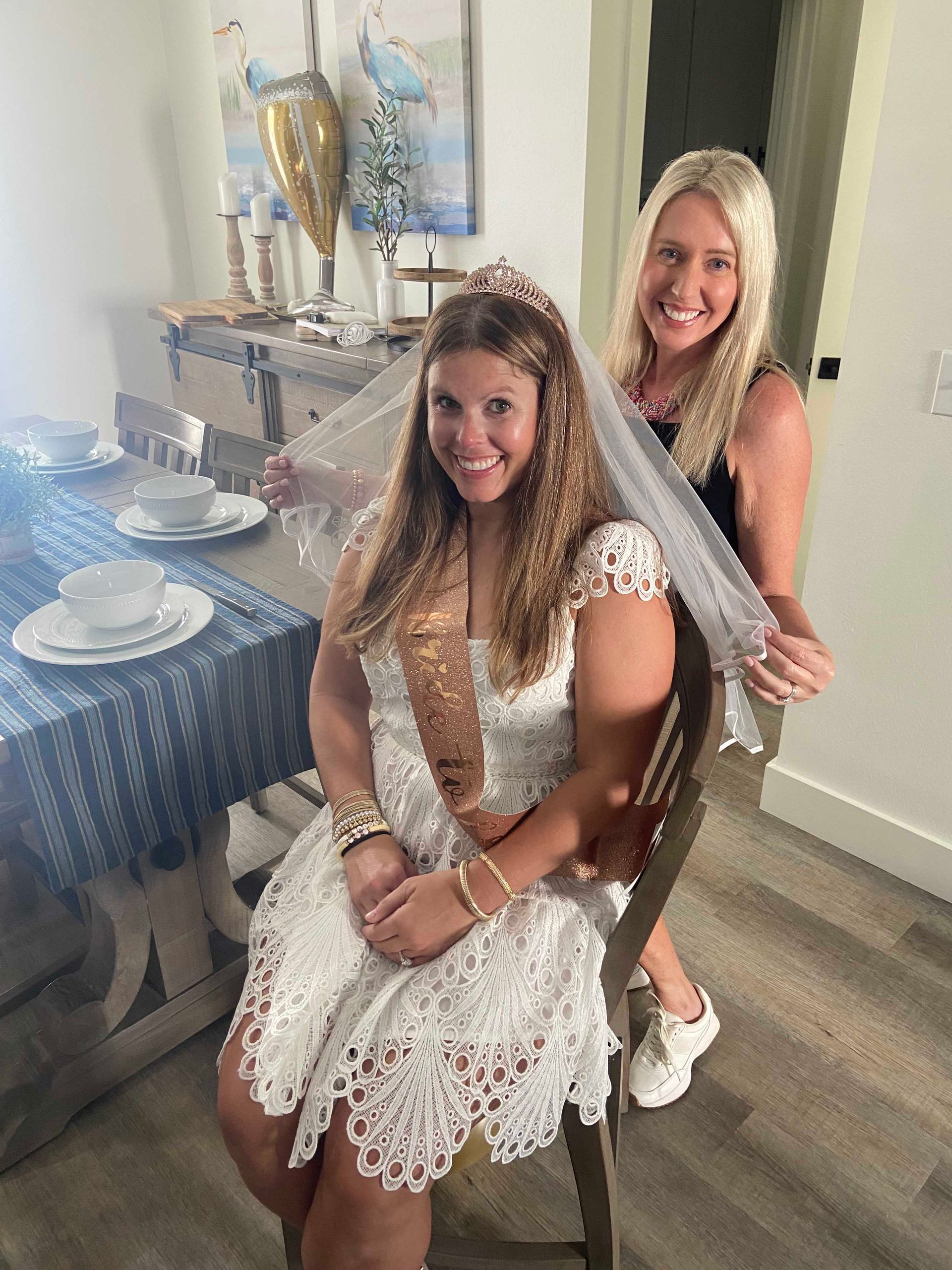 Bride-to-be in white dress with a veil, wearing a tiara and sash, has a friend holding her veil. They are indoors.
