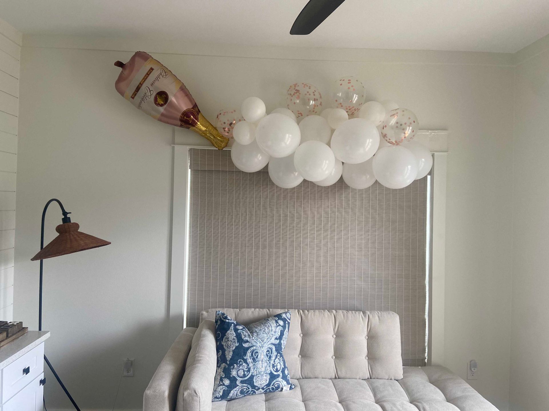 A champagne bottle balloon and white balloons are arranged above a couch in a room with a window.