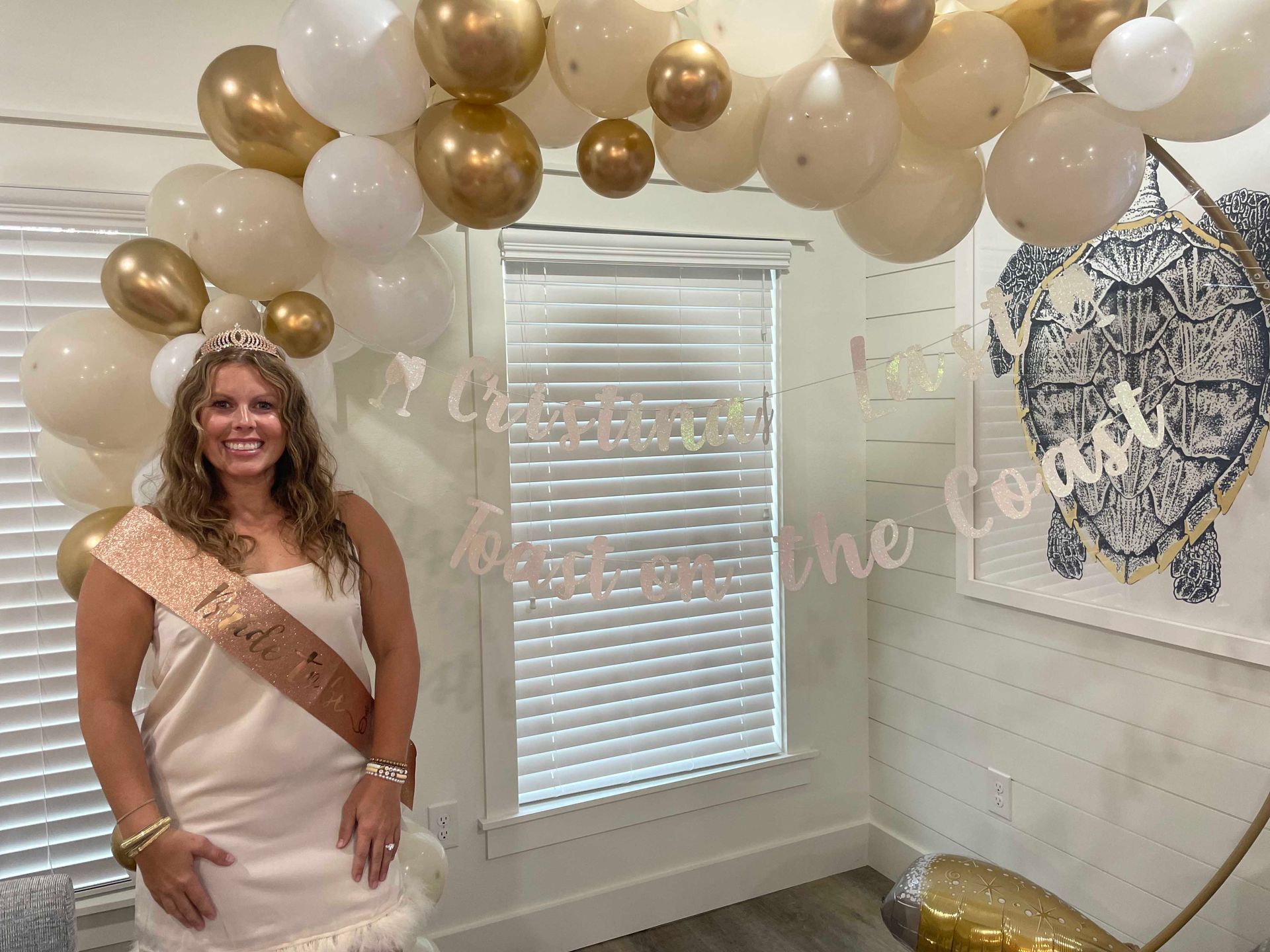 Woman in a white dress and tiara poses under a balloon arch. She wears a sash and smiles in a room with coastal decor.