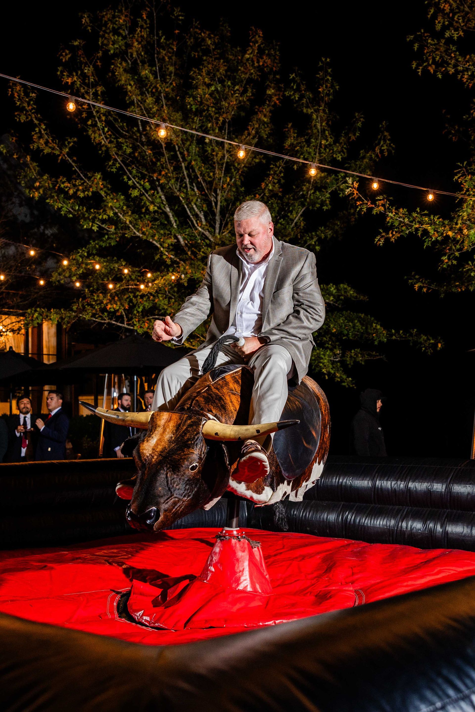 Man in a suit rides a mechanical bull at night. Red mat, twinkling lights in the background.