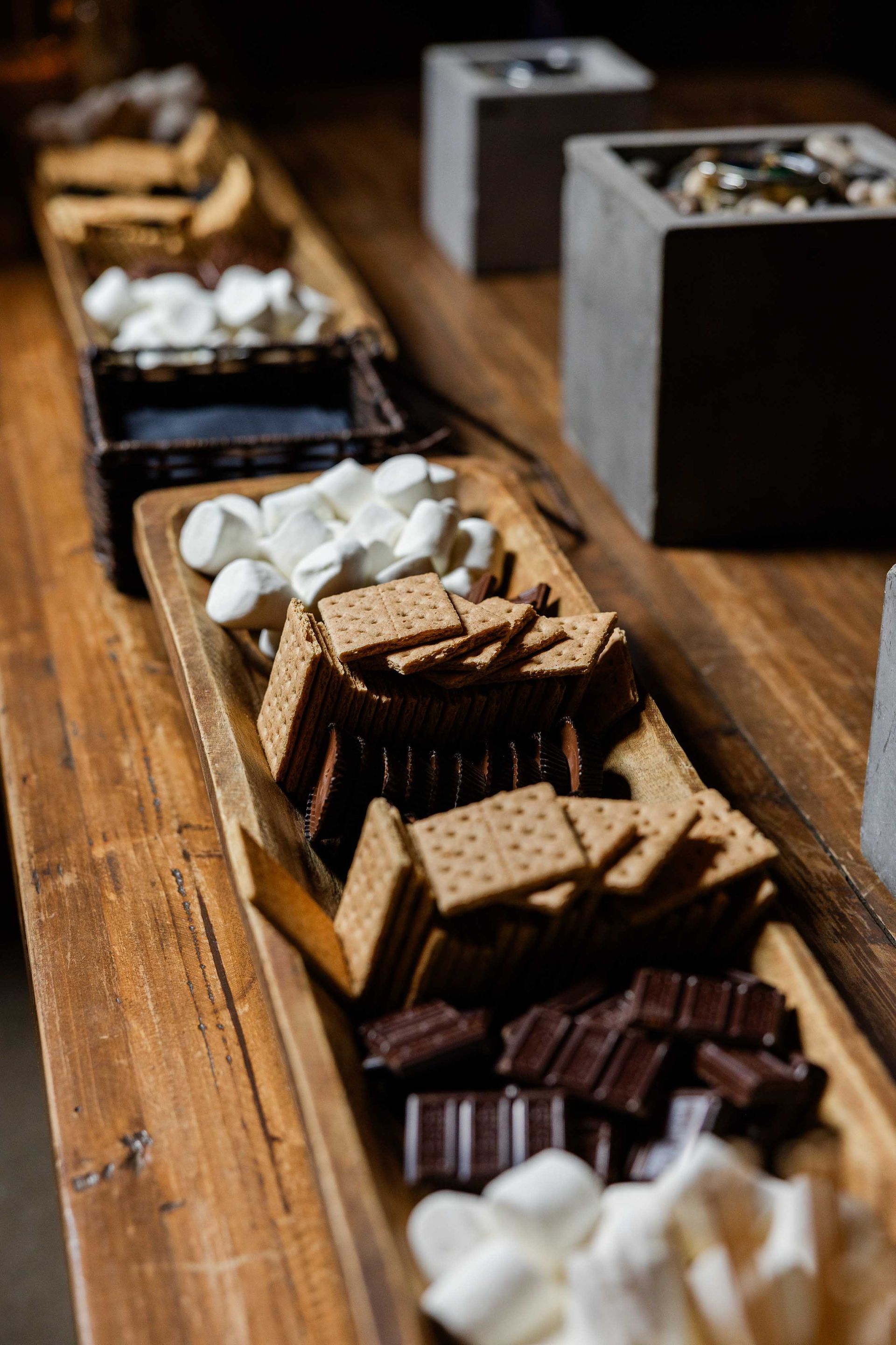 A wooden tray filled with ingredients for s'mores: marshmallows, graham crackers, and chocolate. The tray sits on a wooden surface.