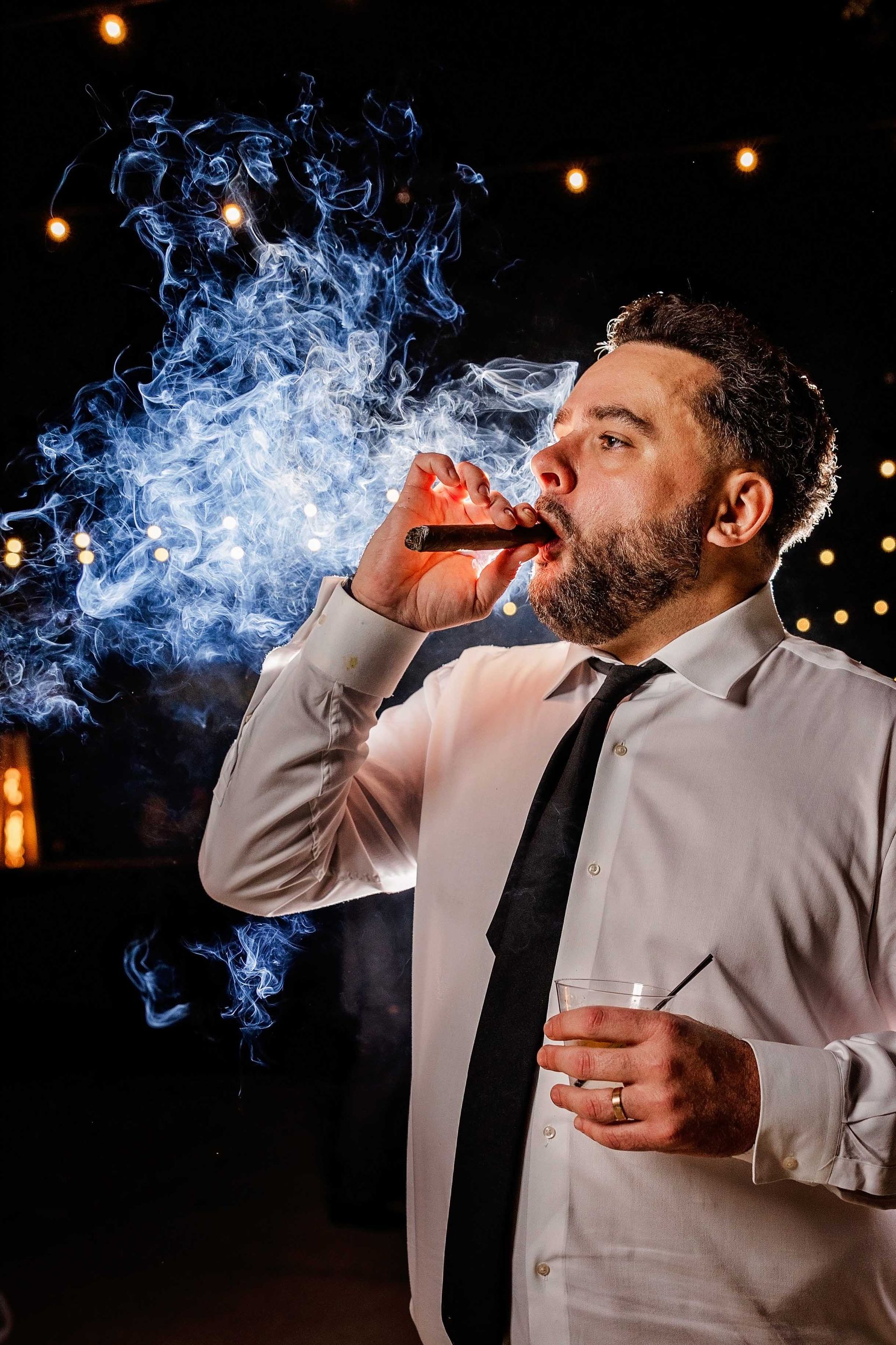 Man in white shirt and black tie exhales cigar smoke, holding a glass of liquid. Dark setting, string lights.