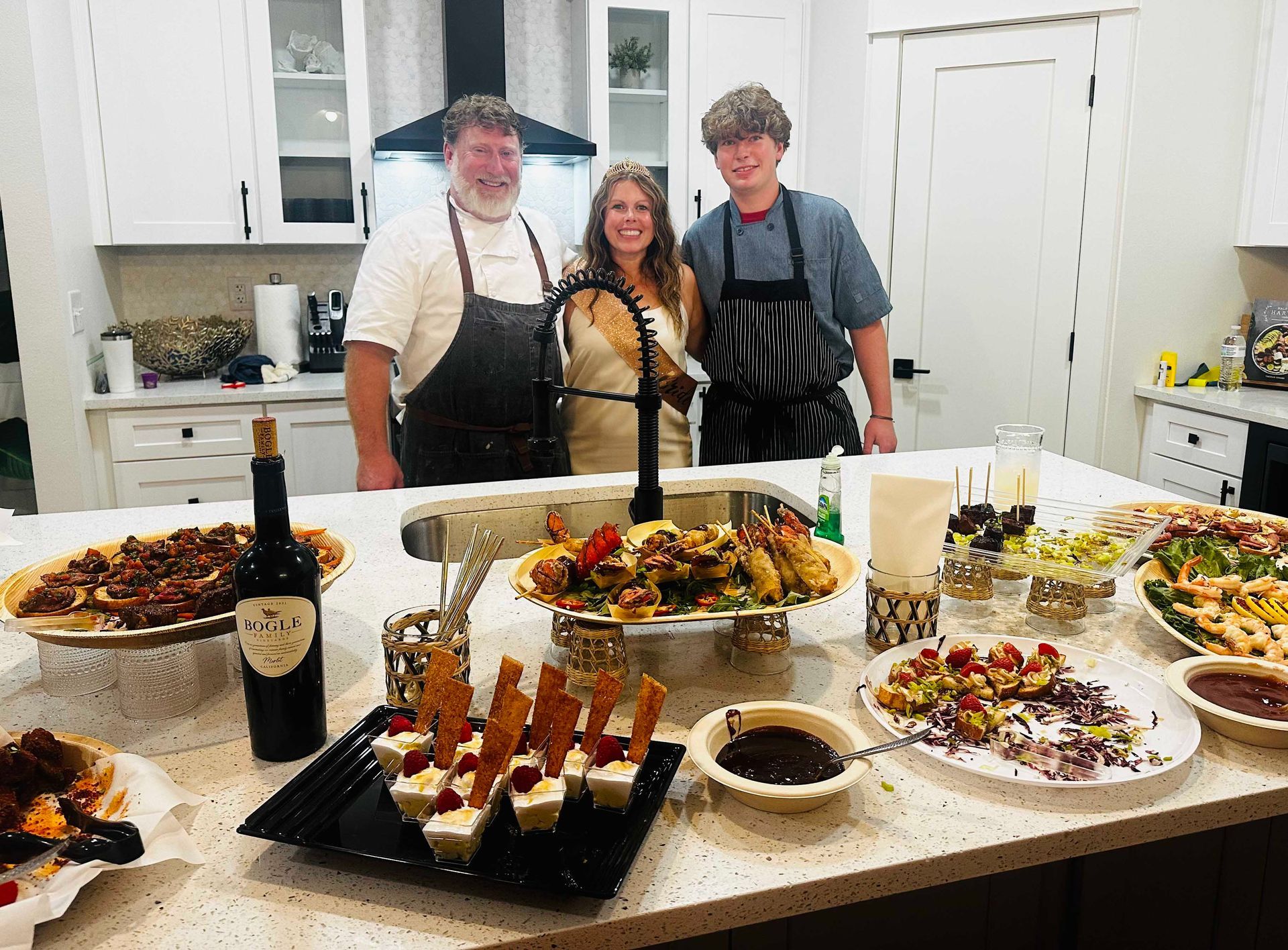 Three people smiling behind a kitchen island laden with food. A man and a teen boy wear chef's aprons; a woman stands in the center.