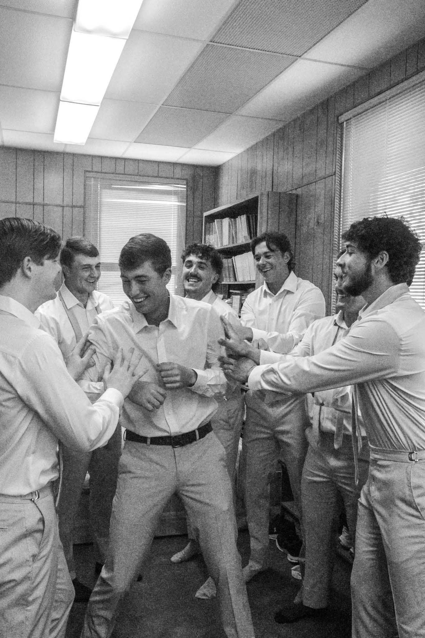 A group of young men in light-colored shirts and pants playfully surround another, who appears to be dancing in an office setting.