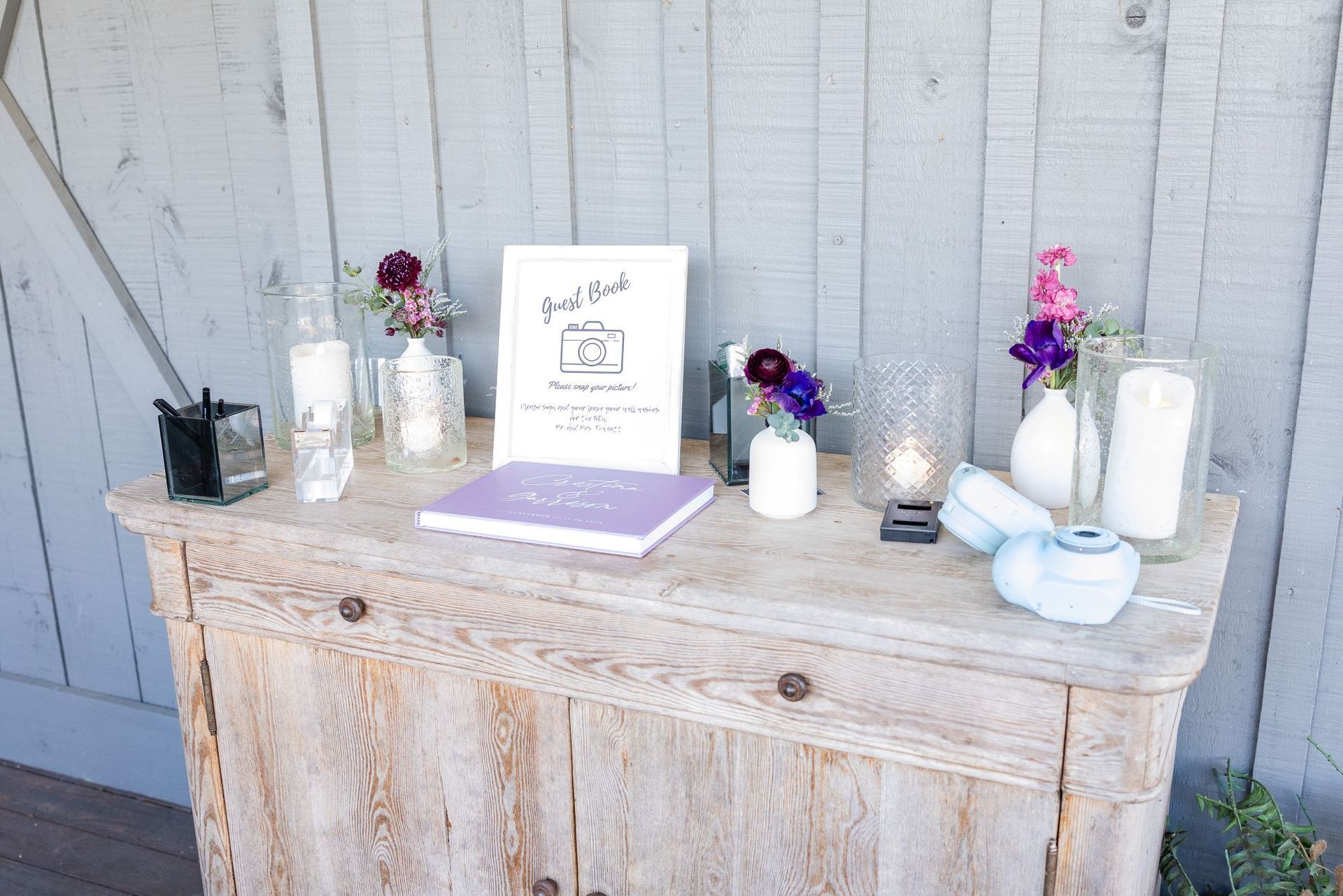 A wooden table decorated with flowers, candles, and a guest book in front of a weathered, gray wall.
