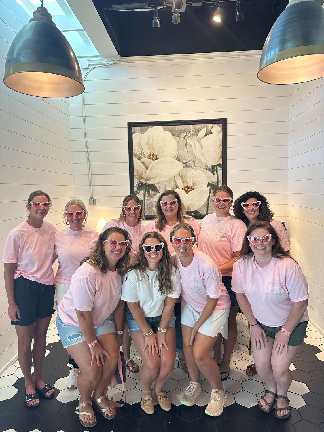 Group of eleven women wearing pink shirts and sunglasses pose inside a photo booth with floral art on the back wall.