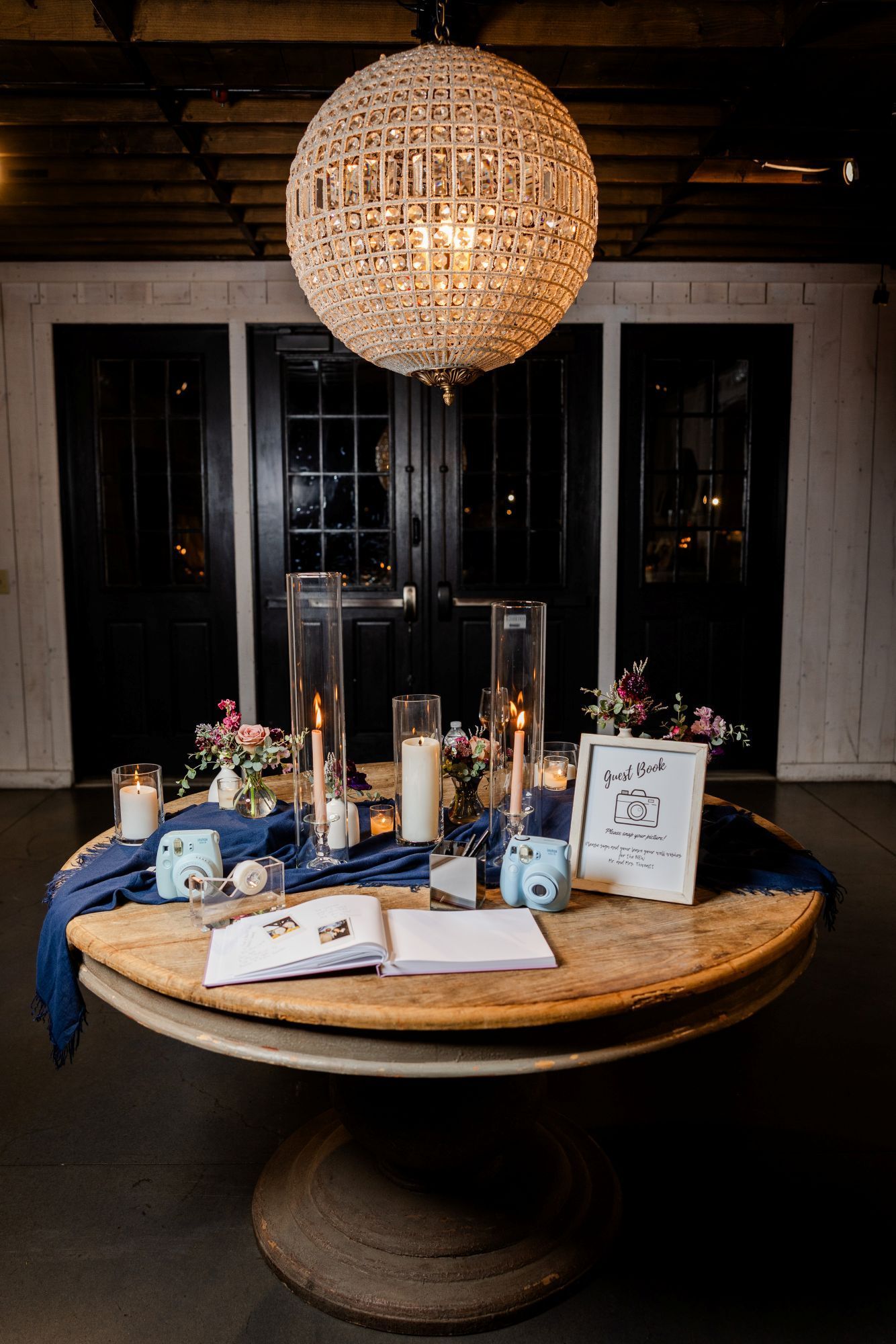 A wedding reception table with candles, flowers, and a guest book, set under a beaded chandelier.