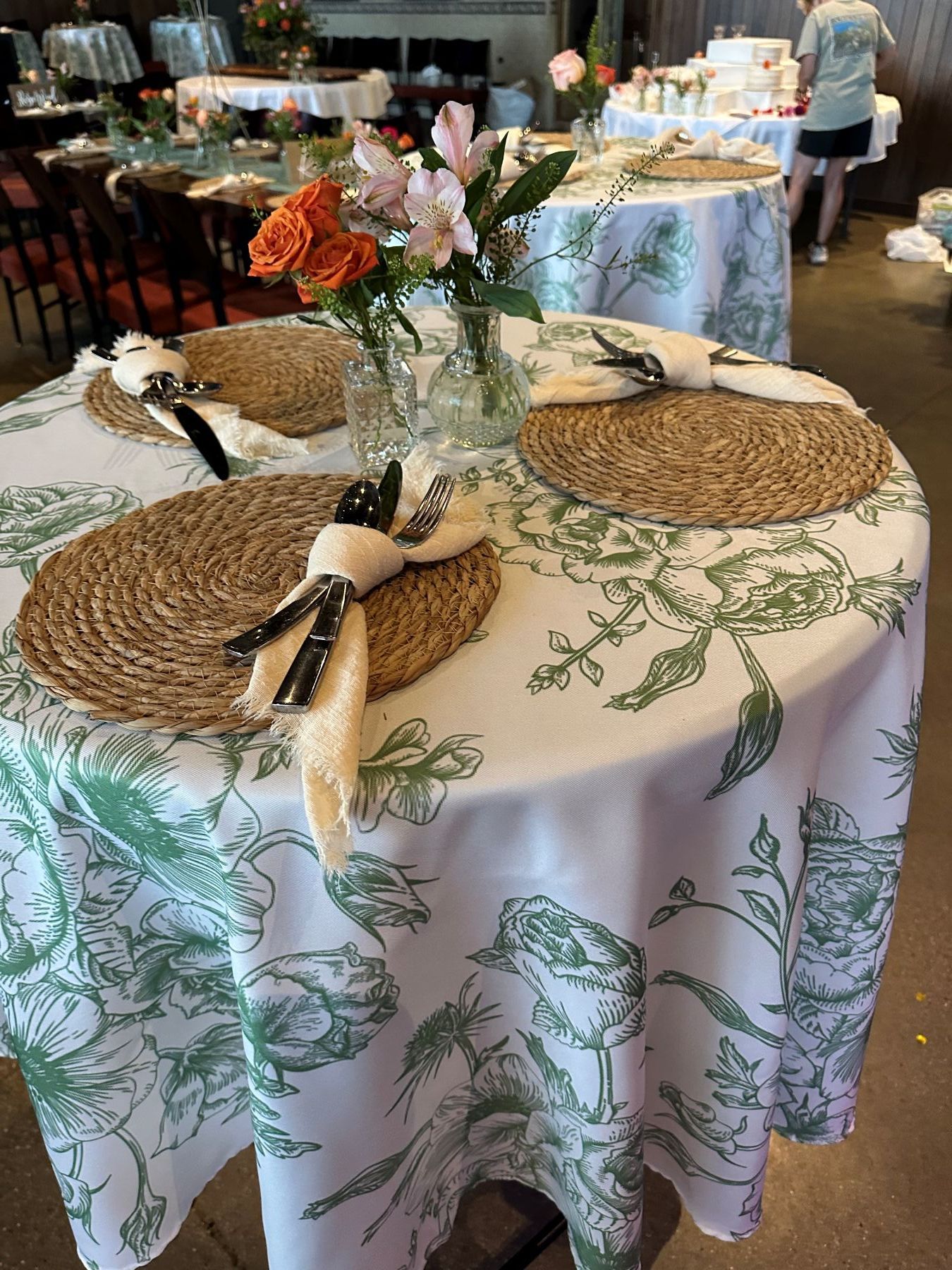 Round table with green floral tablecloth set for dining, featuring woven placemats, napkins, and a flower arrangement.