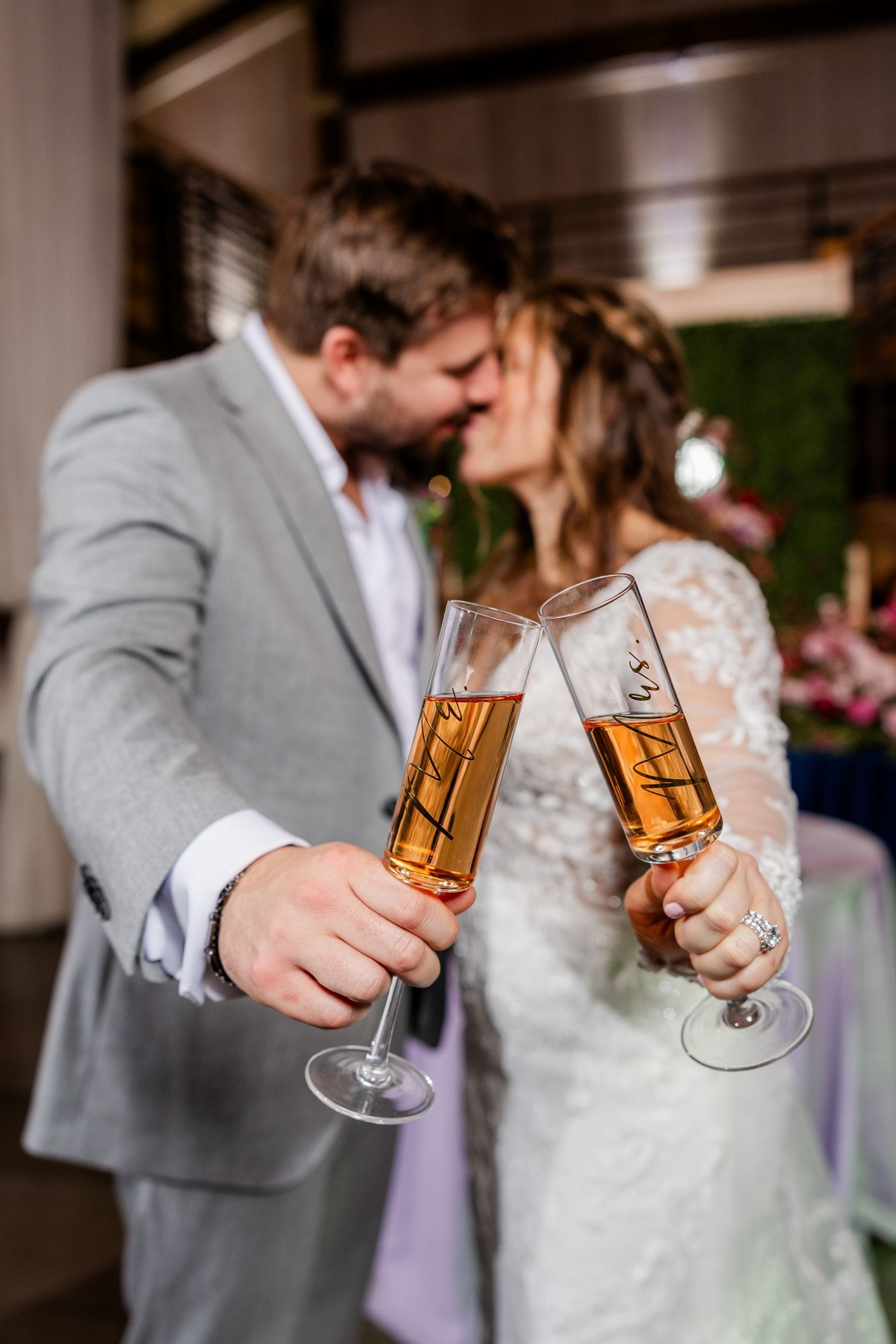 Newlyweds clink champagne glasses while kissing at their wedding reception. The man wears a gray suit, the woman a white lace dress.