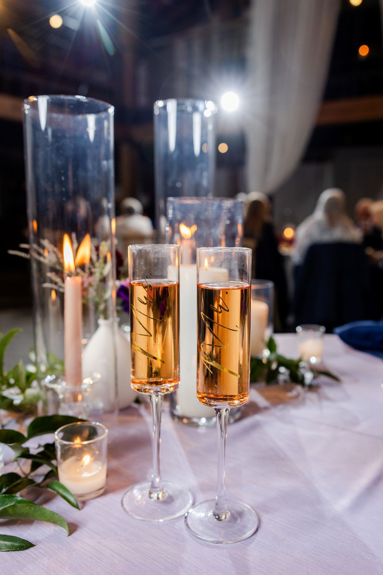 Two champagne flutes with amber liquid on a decorated wedding table, candles and greenery.