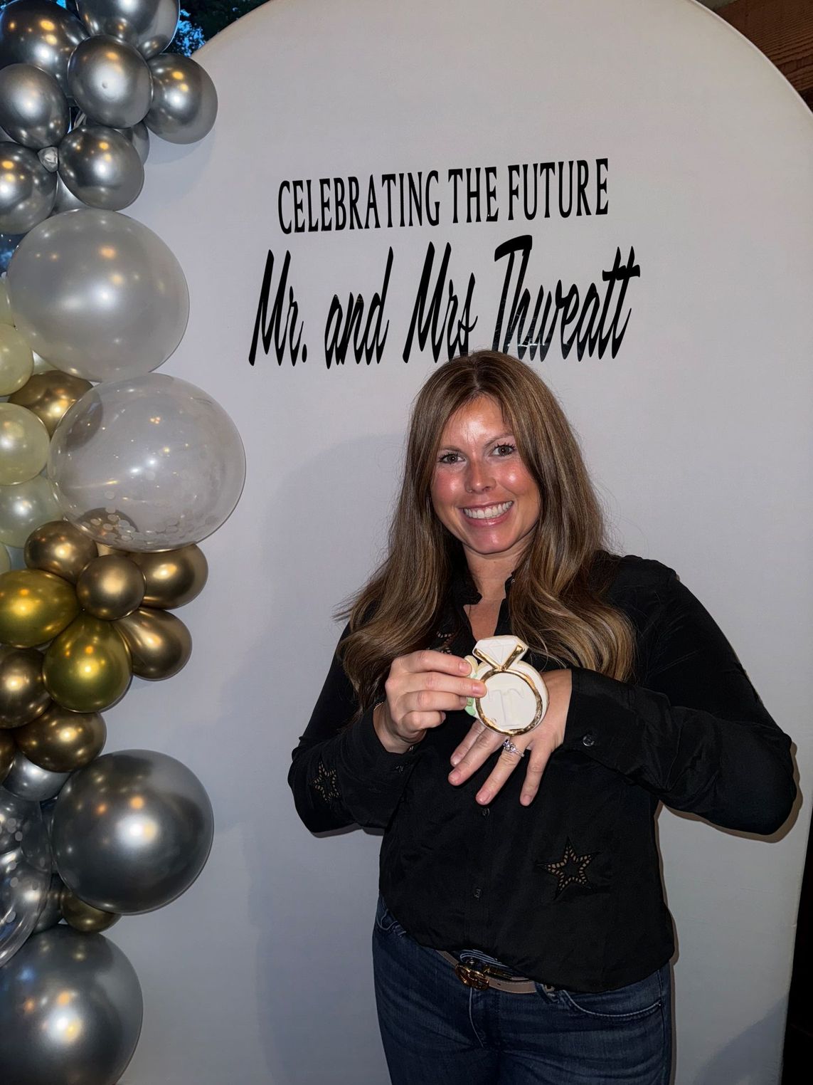 Woman holding a decorative item at an event, smiling. Balloons and a banner that says 