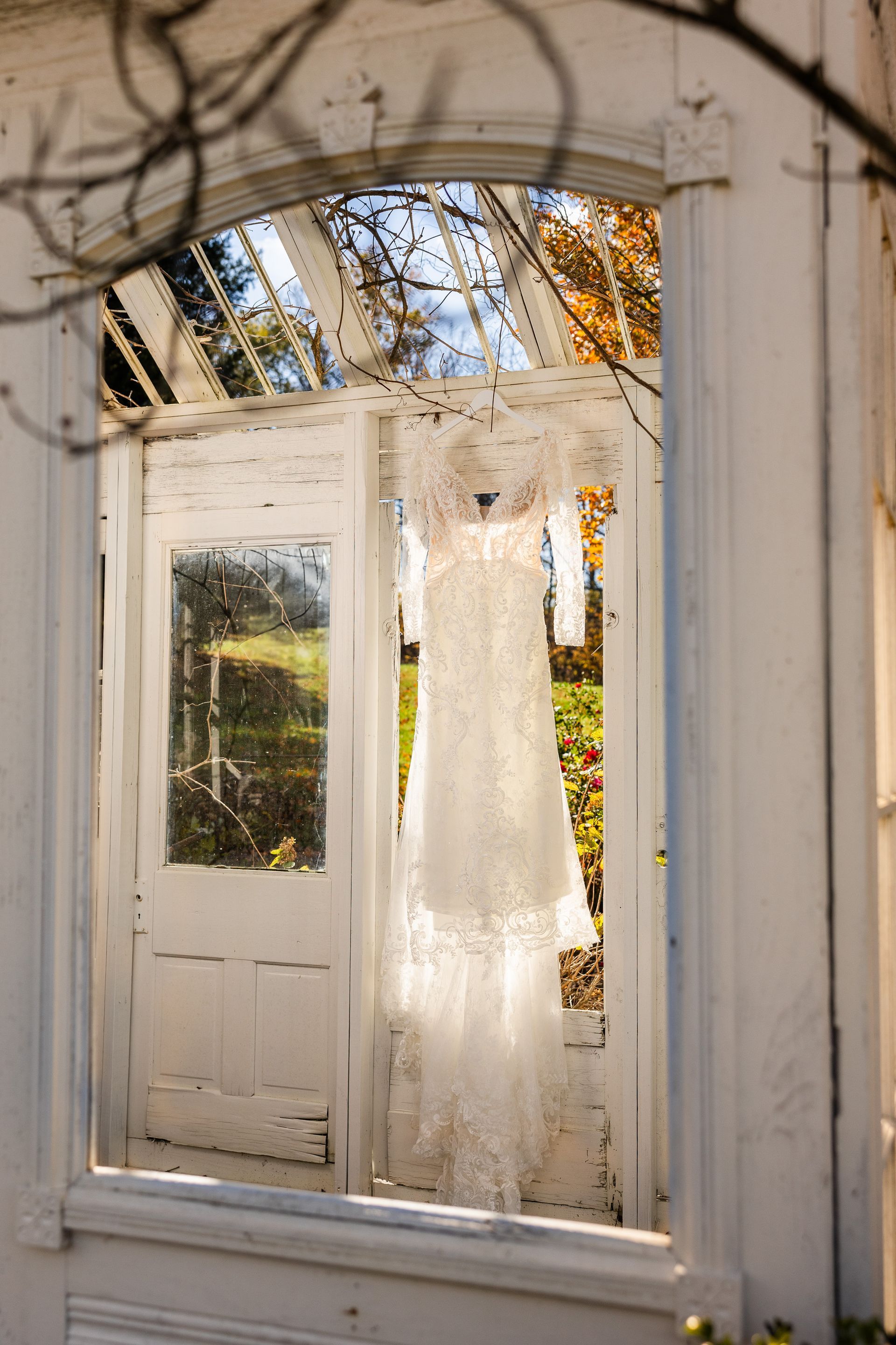 White wedding dress hanging in the doorway of an old, sunlit greenhouse.