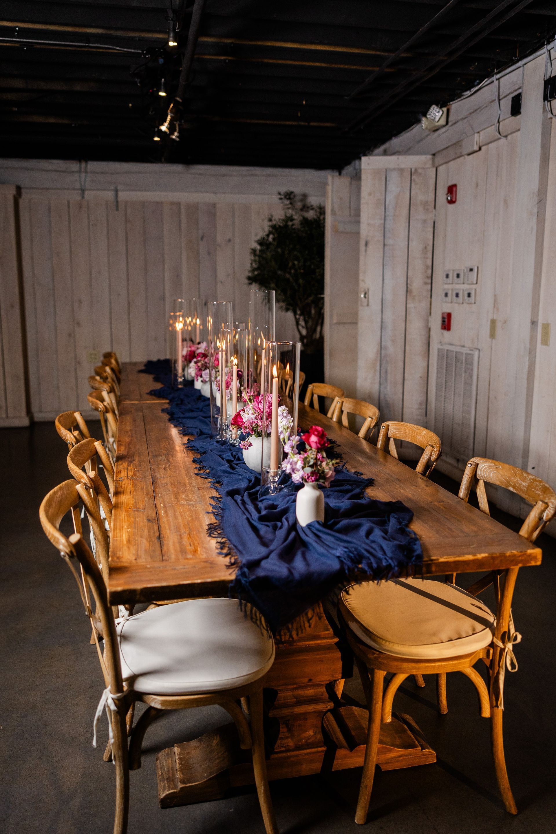 A long wooden table set for a formal event with candles and flowers. Chairs surround the table in an interior with wooden walls.