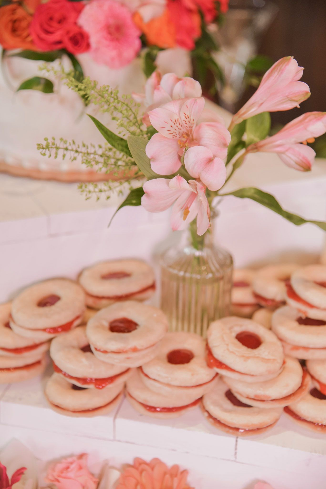Cookies with red filling are stacked on a table with pink flowers in a vase, and a cake with pink and orange flowers.