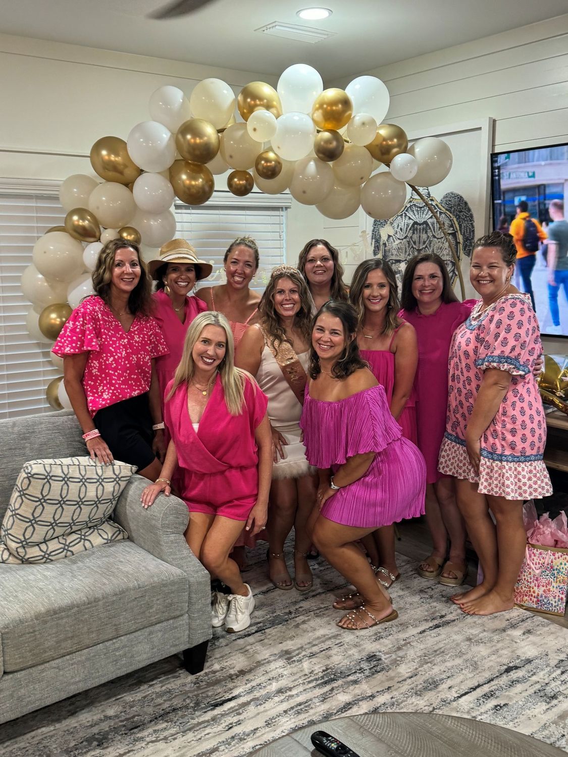 Group of women in pink outfits pose in front of a balloon arch. They are indoors, possibly at a celebration, with the women smiling.