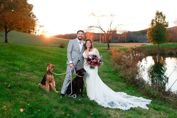 Newlyweds with two dogs pose by a pond, the sun setting on a green field.