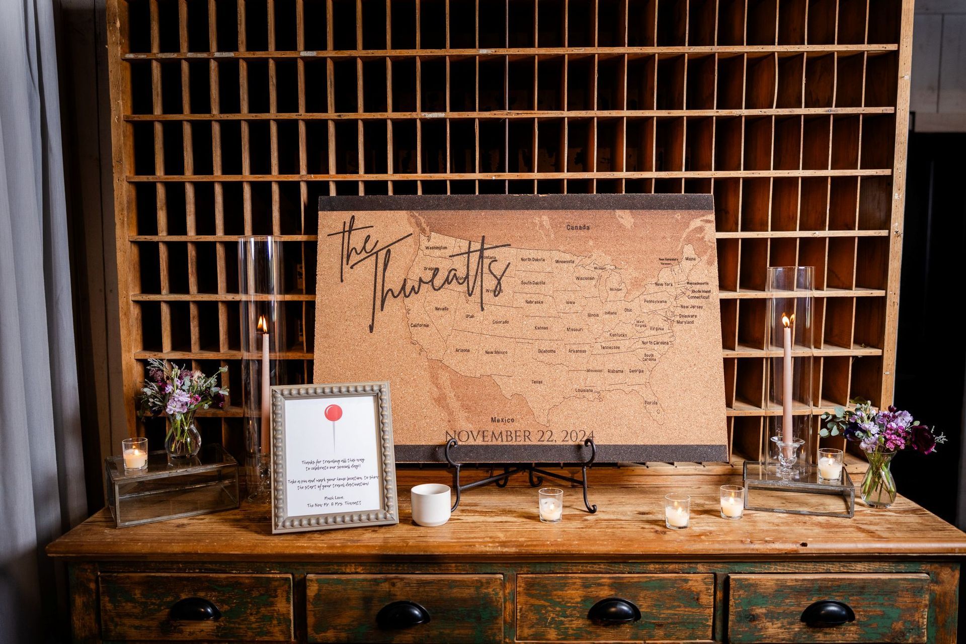 A rustic wedding reception table features a cork board guestbook map, framed sign, candles, and a wooden cubby shelf backdrop.