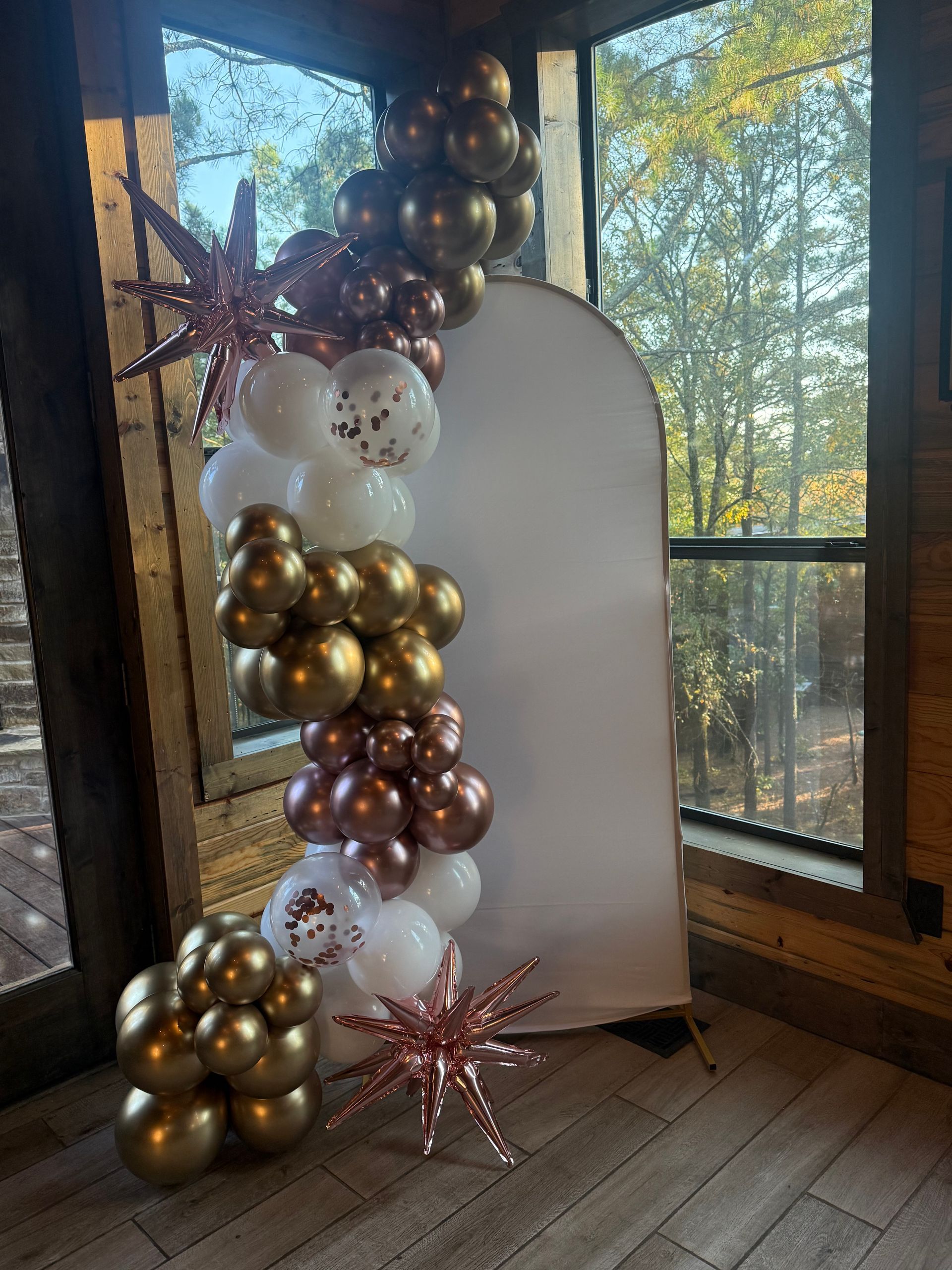 An arched white backdrop decorated with a cascading balloon garland in gold, rose gold, and white, with star accents.