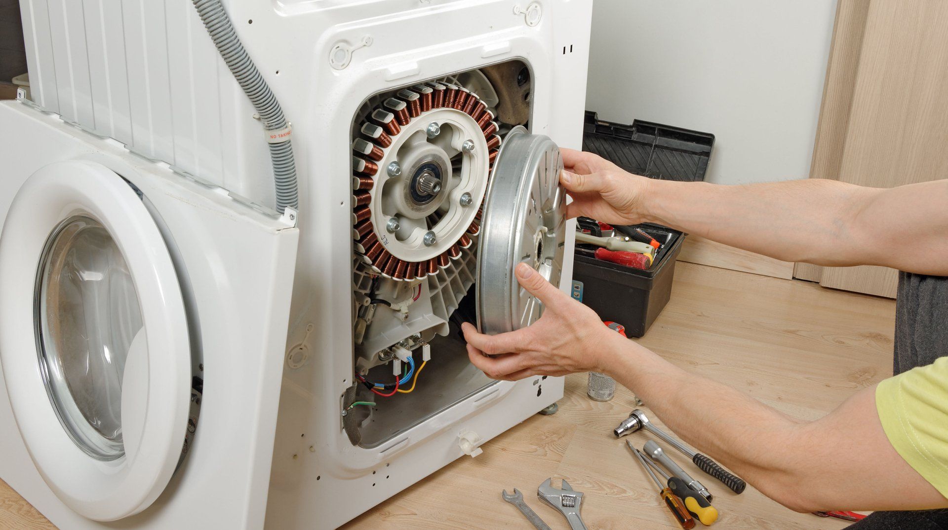 Person repairing washing machine, holding a large metal part near the motor; tools visible.