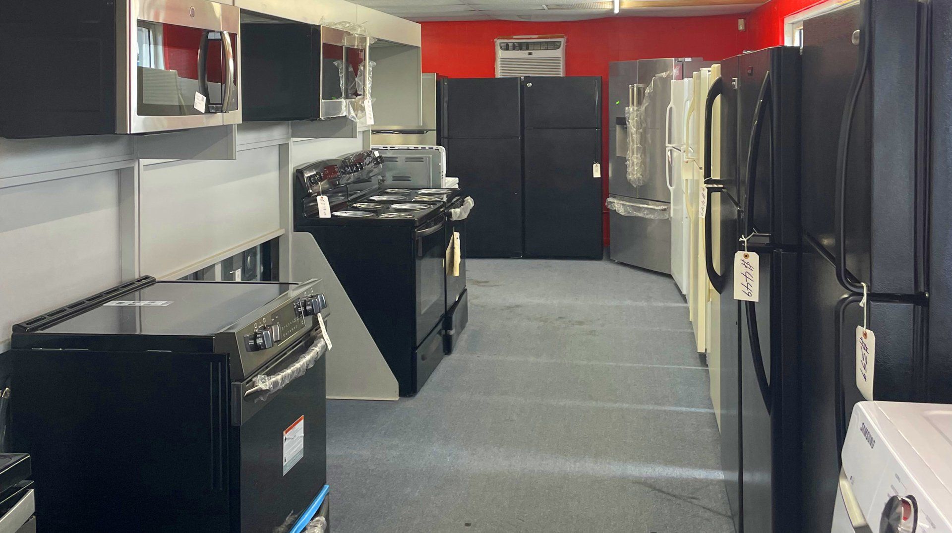 Appliances in a showroom: black and silver refrigerators, stoves, and microwave against red walls.