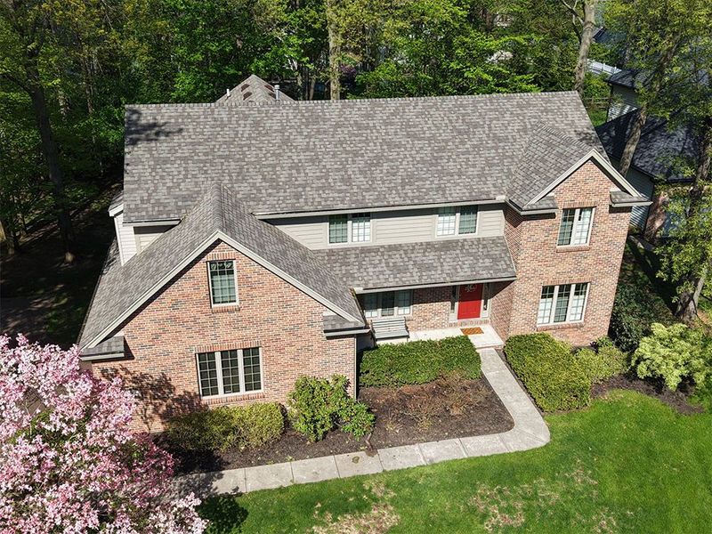 Aerial view of a brick suburban house with gray roof, front walk, and green lawn surrounded by trees
