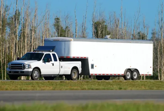 A white pickup truck towing a large white enclosed trailer along a highway with a forest in the background.