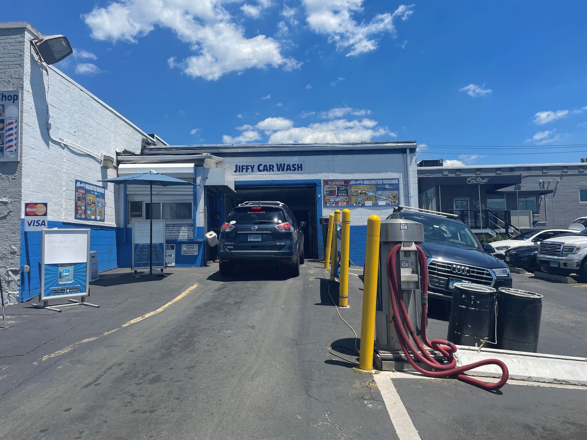 Car entering a blue and white car wash on a sunny day. A vacuum and a few other cars are present.