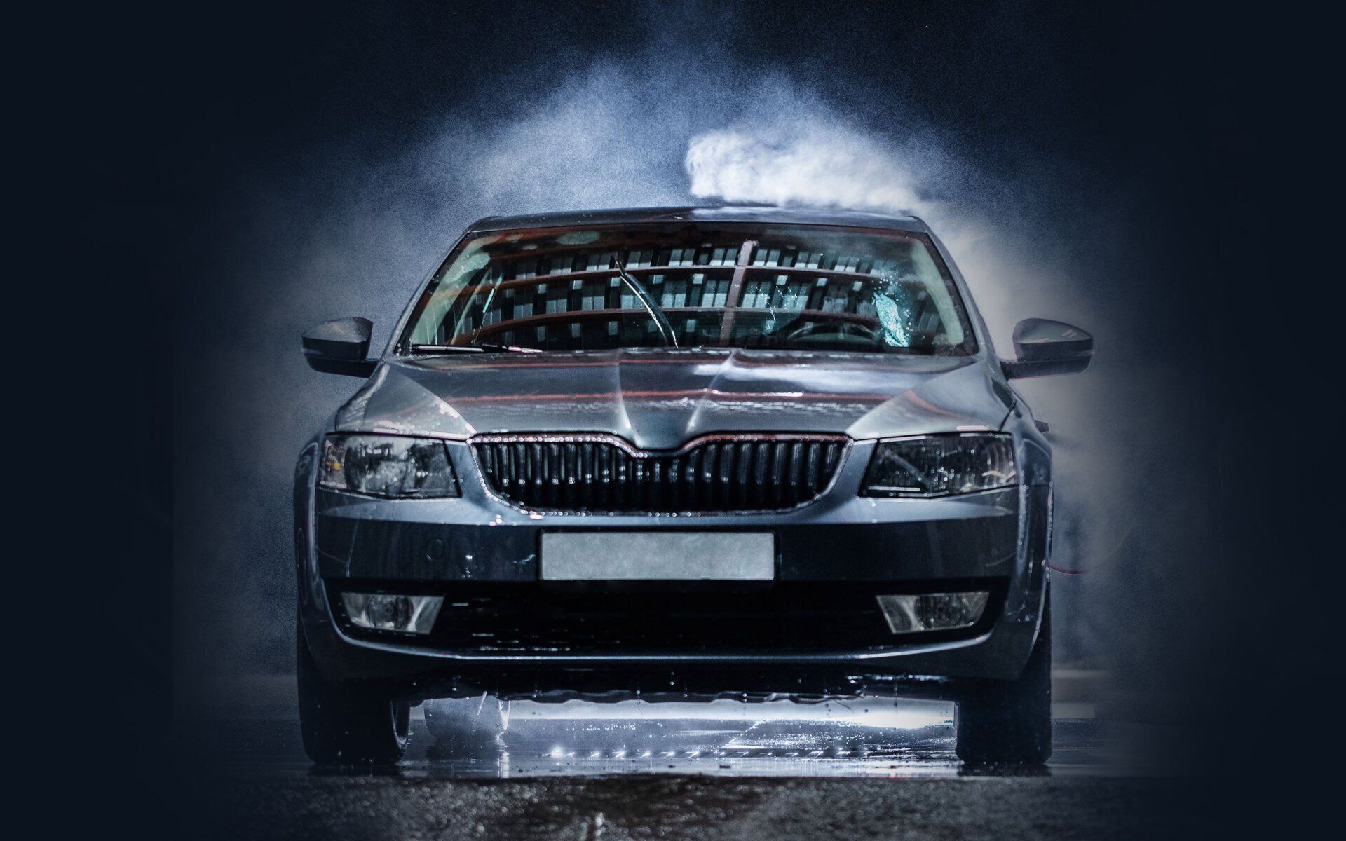 A dark gray car being washed with water spray, front view, in a dark setting.