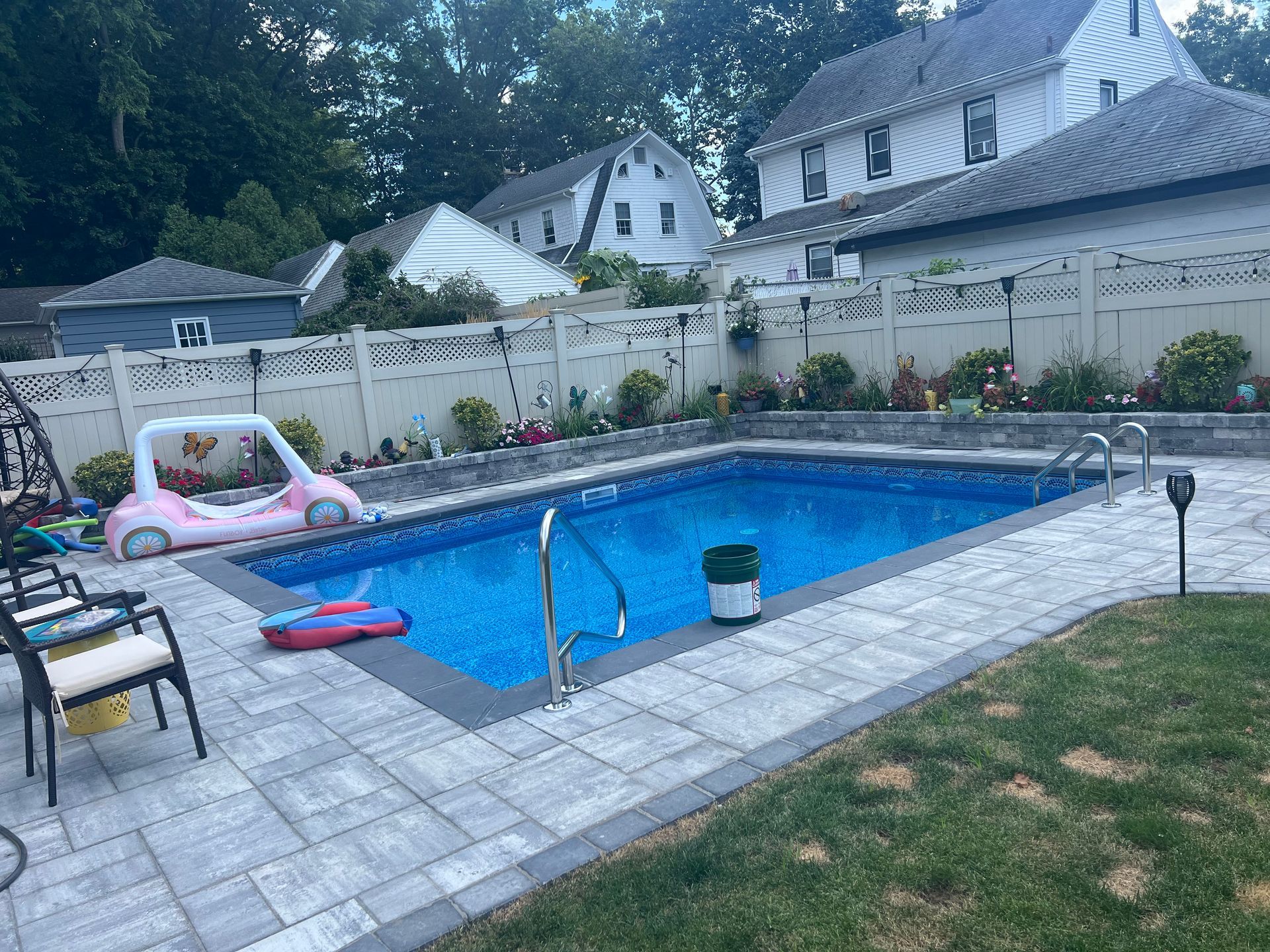 Backyard swimming pool with blue water, stone patio, pink float, and a fenced yard with houses behind it