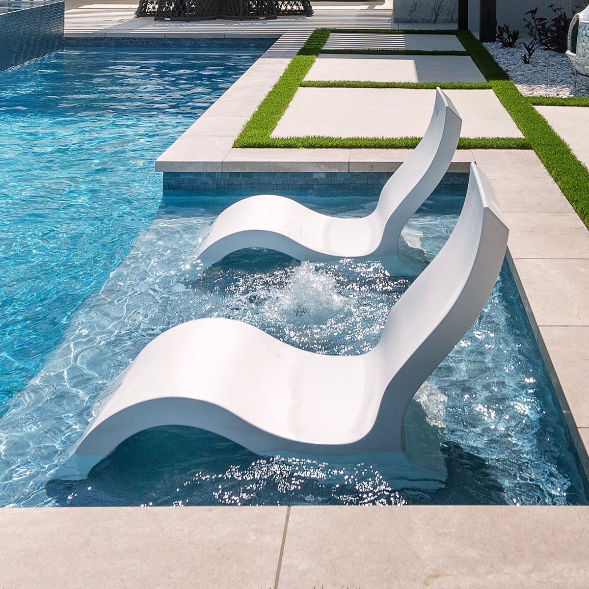 White sculptural lounge chairs partially submerged in a blue swimming pool beside a modern patio.