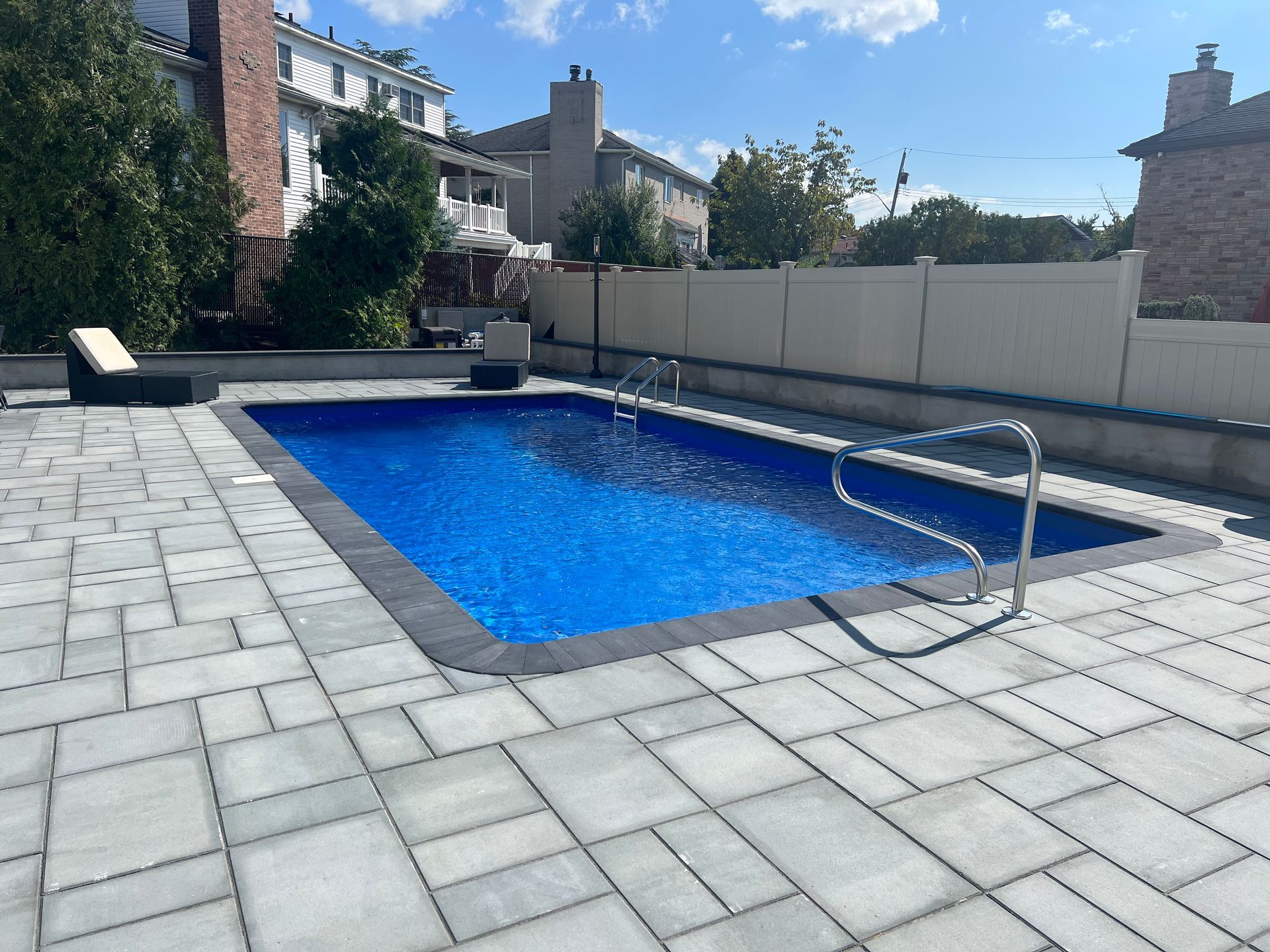 Backyard swimming pool with blue water, stone deck, and a metal handrail under a sunny sky