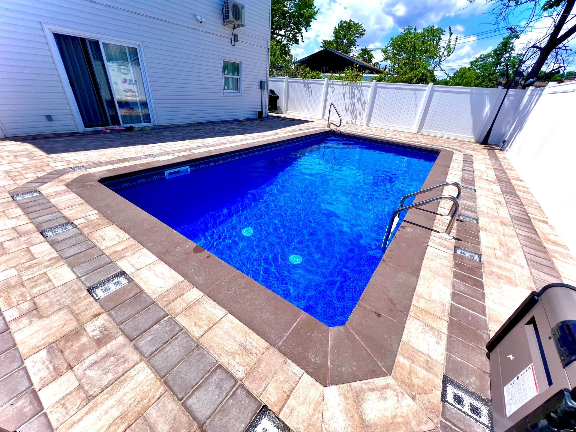 Backyard with a blue rectangular swimming pool, tan paver deck, and white privacy fence under a sunny sky