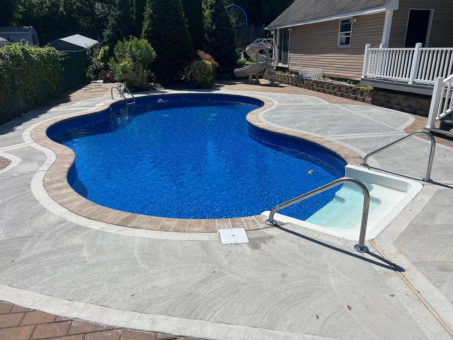 Curved backyard swimming pool with blue water, surrounding patio, and steps with handrail