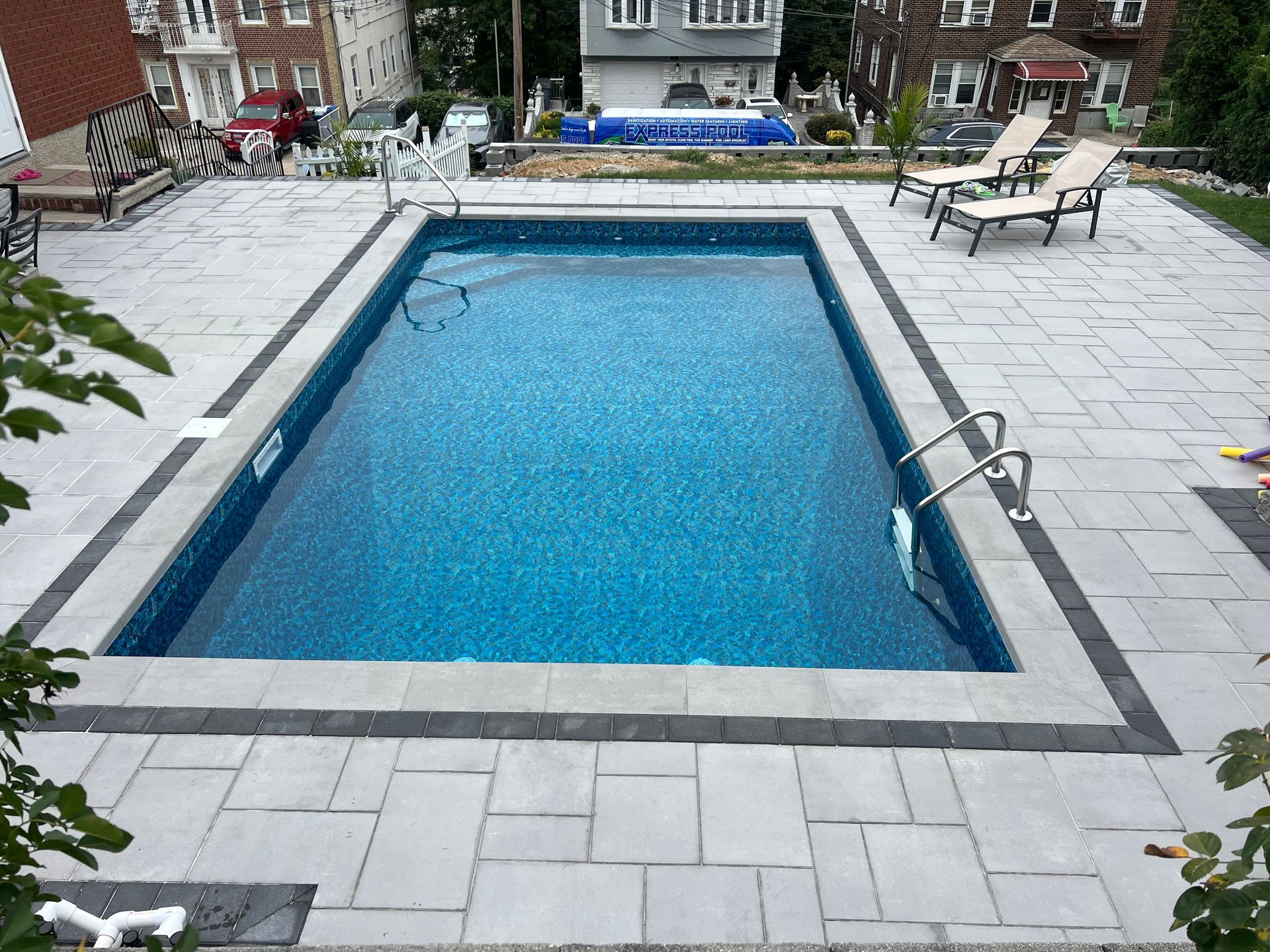 Rectangular outdoor swimming pool with blue water, surrounded by gray stone patio and lounge chairs.