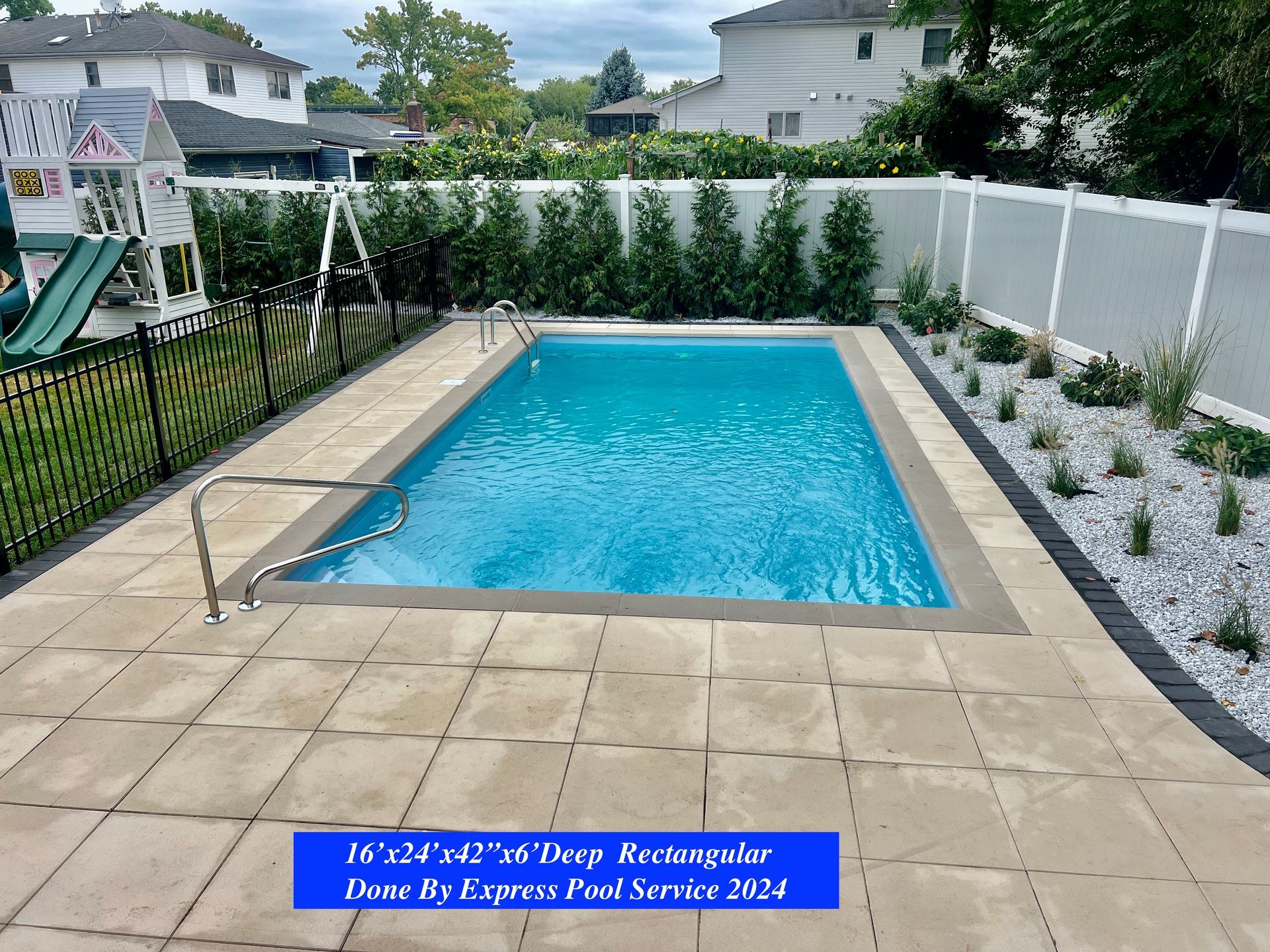 Backyard swimming pool with stone patio, white fence, and landscaped plants