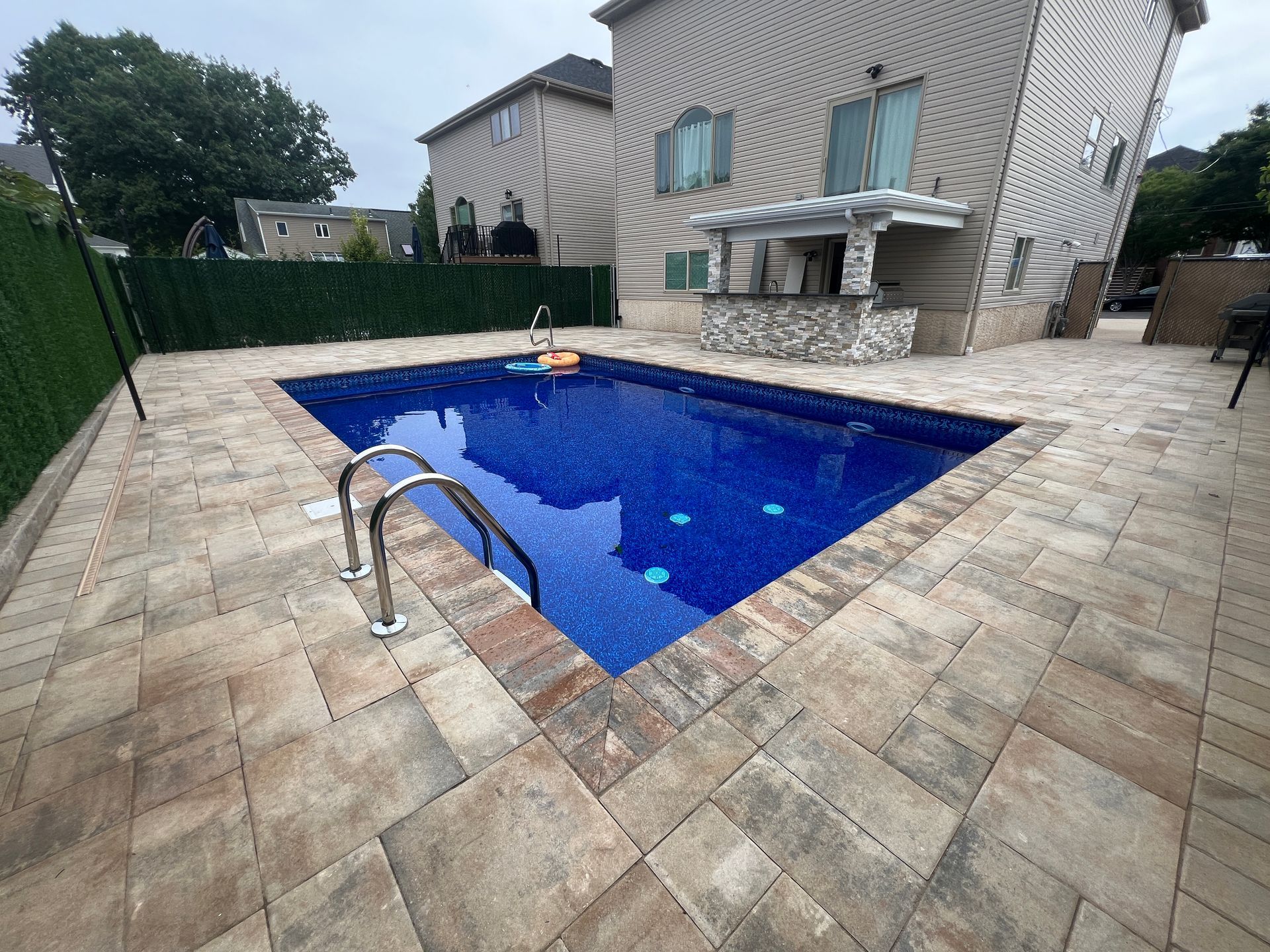 Backyard patio with a blue in-ground pool beside a house and stone paving
