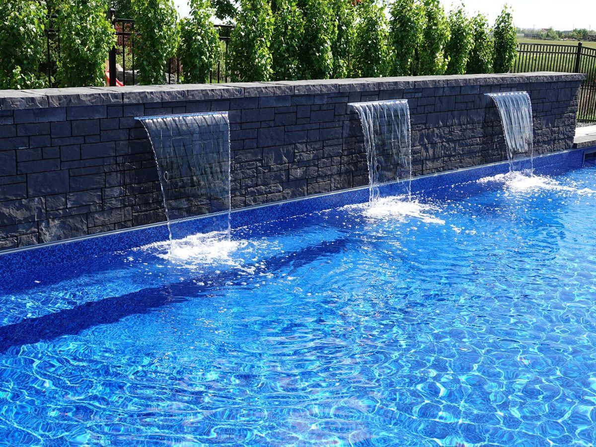 Blue swimming pool with water fountains beside a dark stone wall and green shrubs