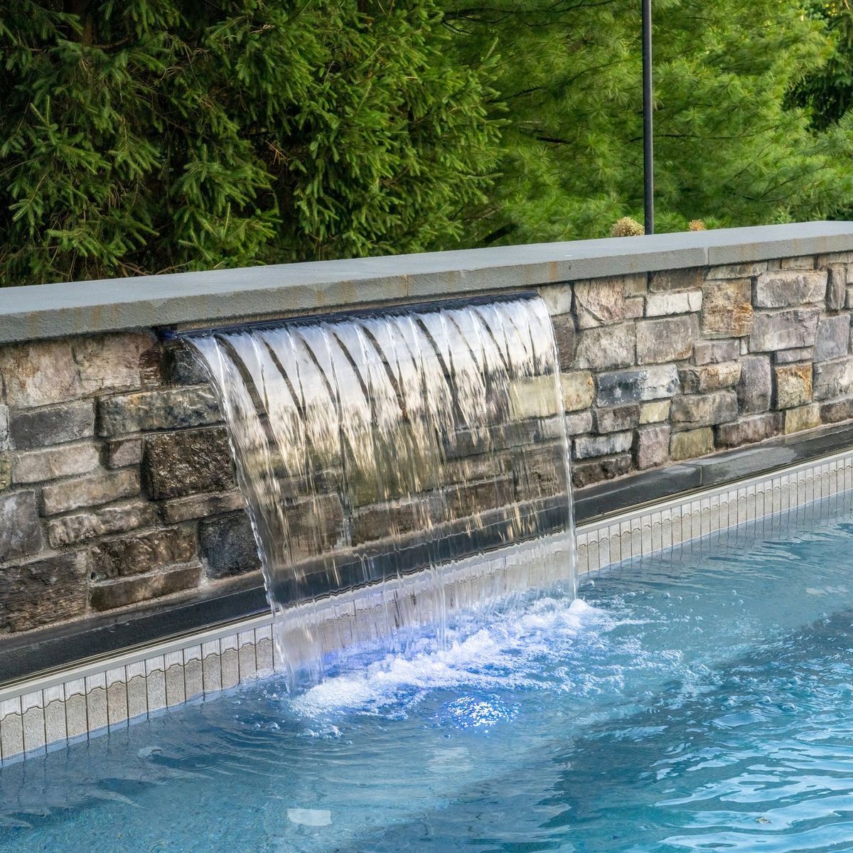 Stone wall fountain spilling water into a blue pool beside lush green plants.