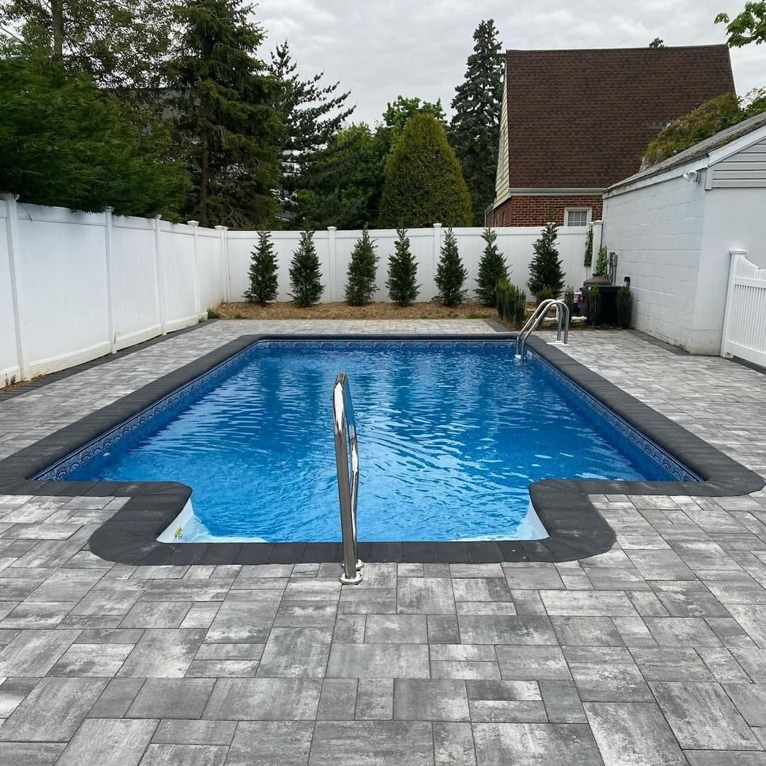 Backyard swimming pool with gray stone patio, white fence, and evergreen shrubs.