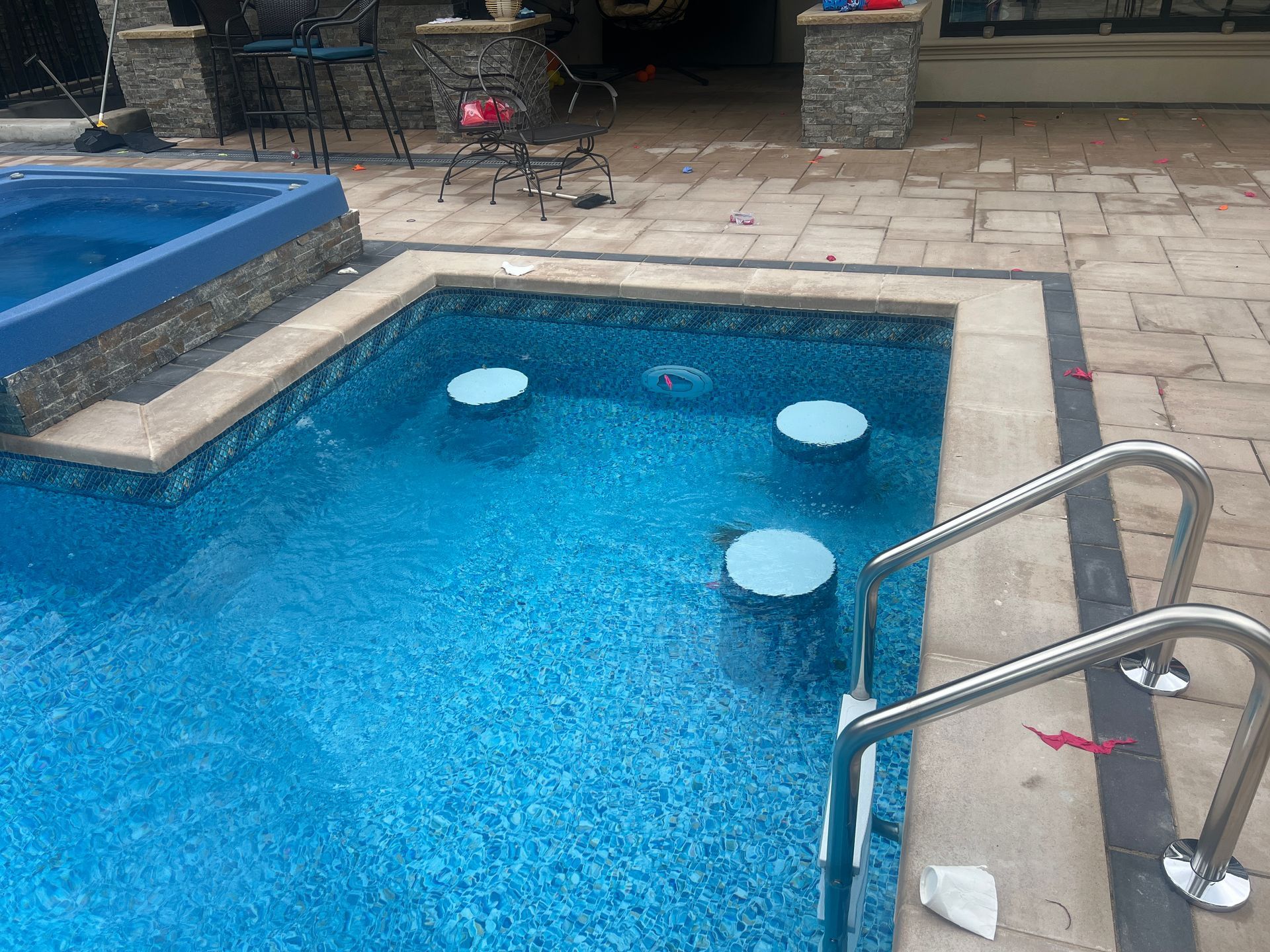 Small backyard pool with blue water, white floating pads, and stone patio steps with a metal handrail