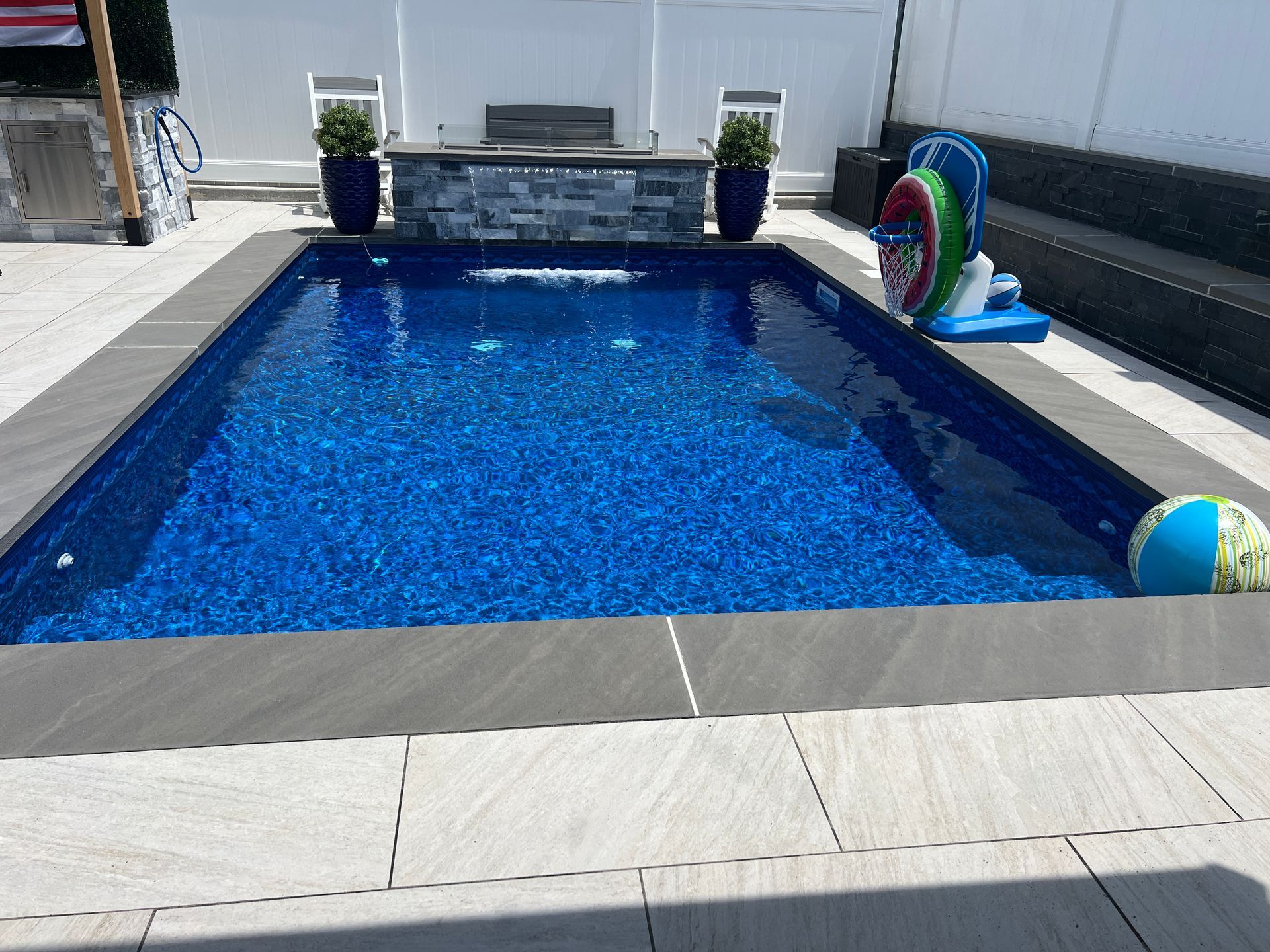Blue backyard swimming pool with a diving board, two potted plants, and pool floats near a white fence