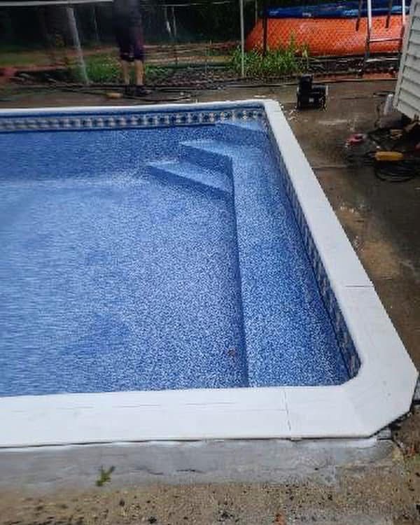 Rectangular in-ground pool with blue mosaic tile and white coping, viewed from a wet backyard patio.
