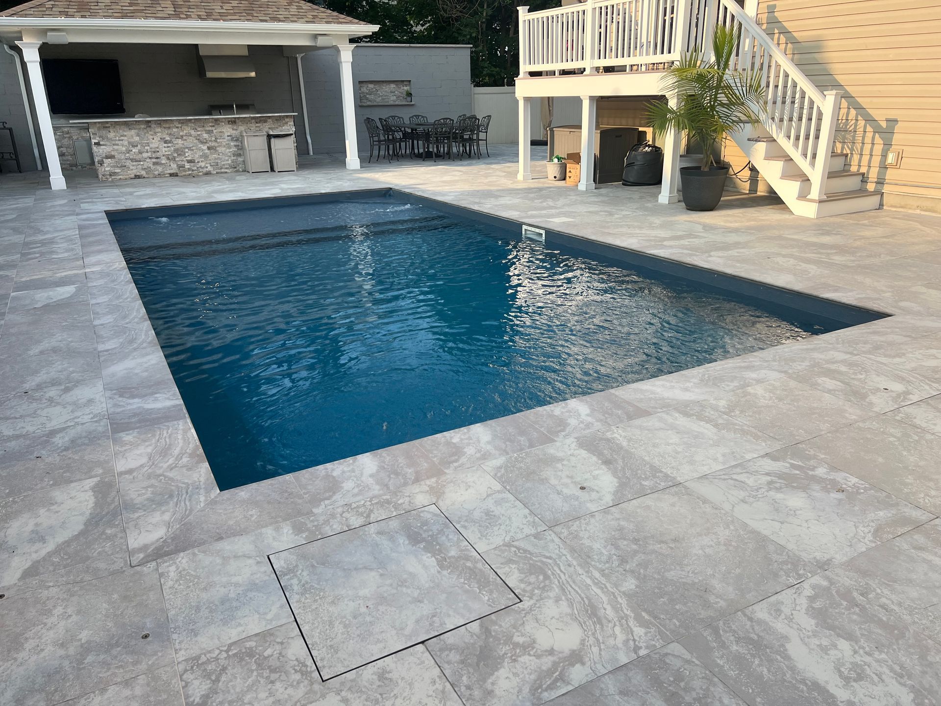 Backyard pool with blue water, light stone deck, patio seating, and stairs to an elevated deck