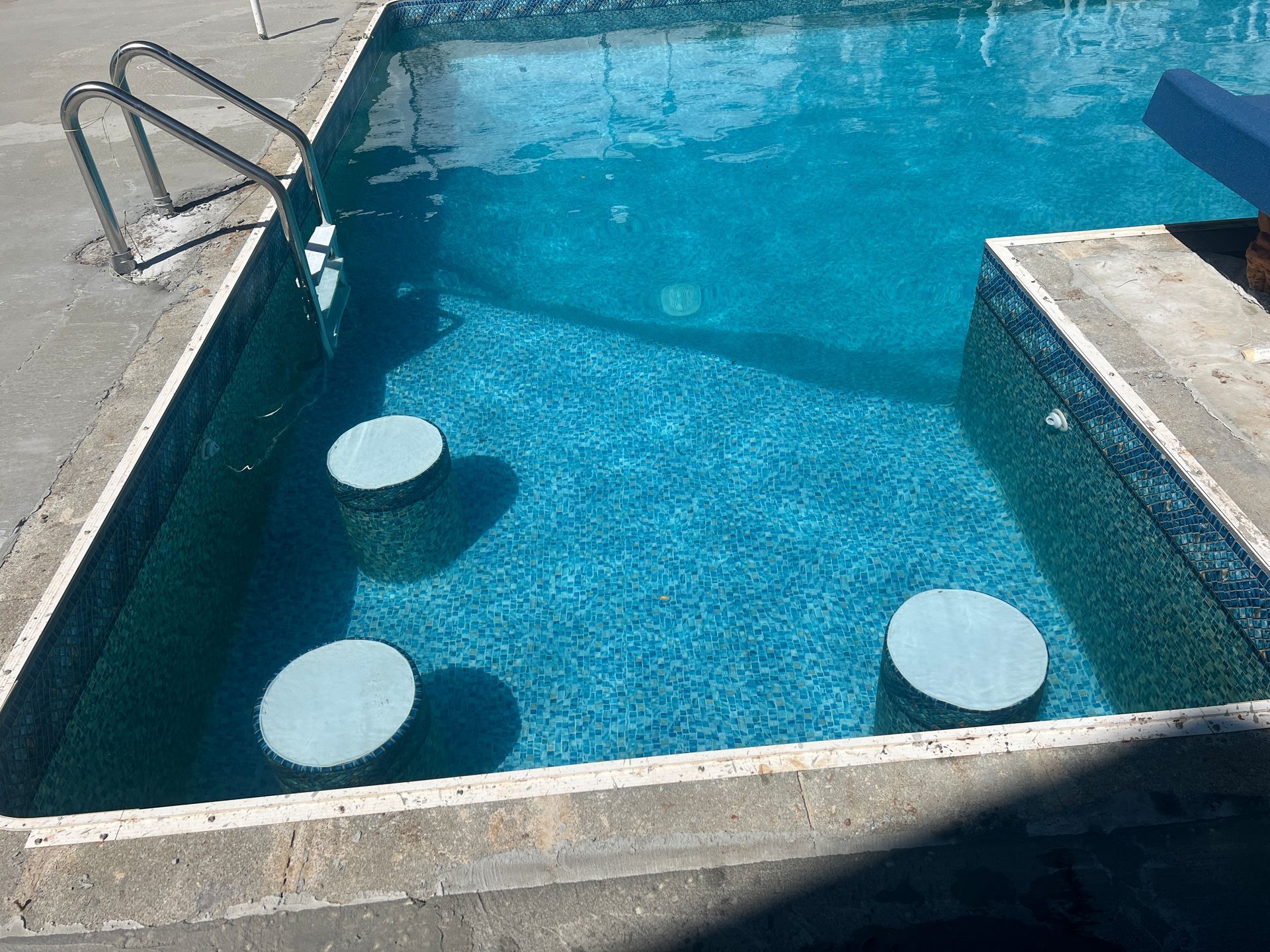 Swimming pool with clear blue water, three round submerged stools, and metal handrails at the edge.
