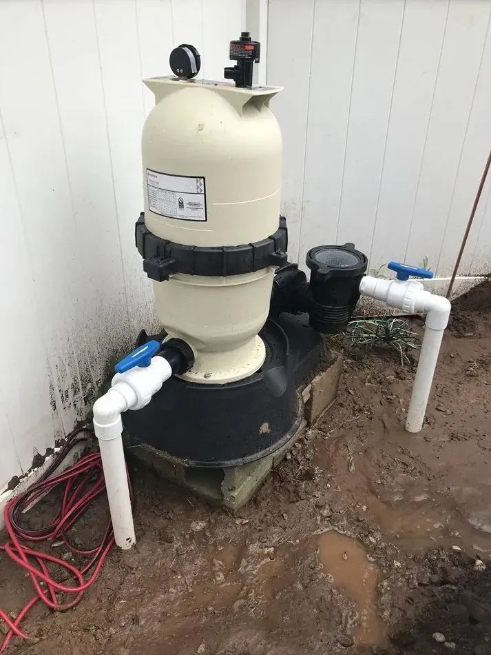Outdoor pool filter and pump system beside a white wall, with pipes and a blue valve in a dirt area.