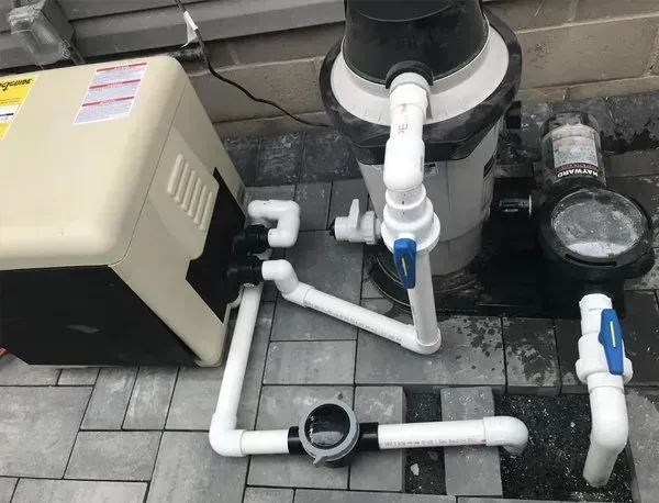 Pool pump and filter plumbing with white PVC pipes and beige equipment on a gray paver patio.