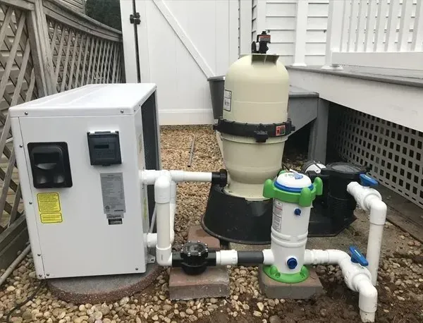 Outdoor pool equipment: white control box, filter tank, and connected pipes beside a house.
