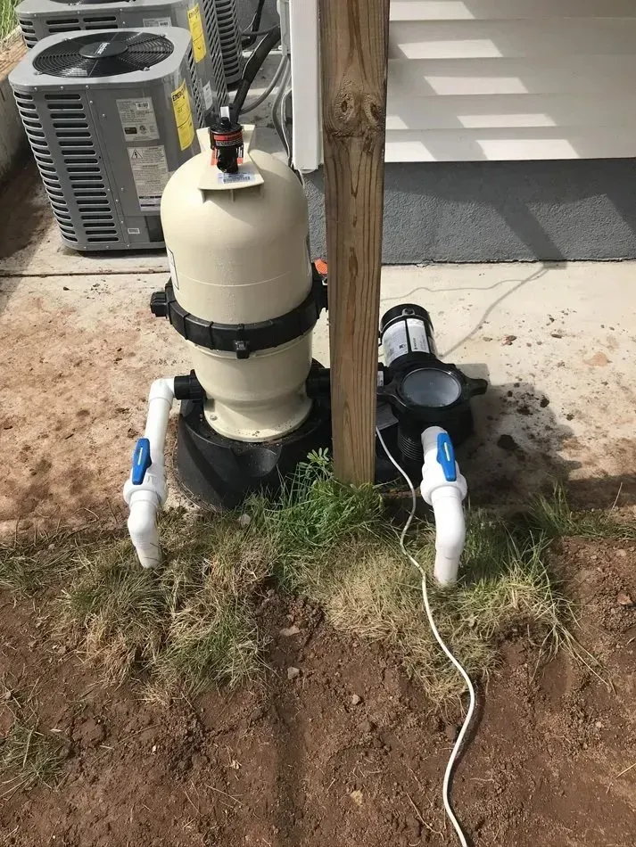 Pool pump and filter equipment beside a wooden post, with hoses and a cord on the ground.